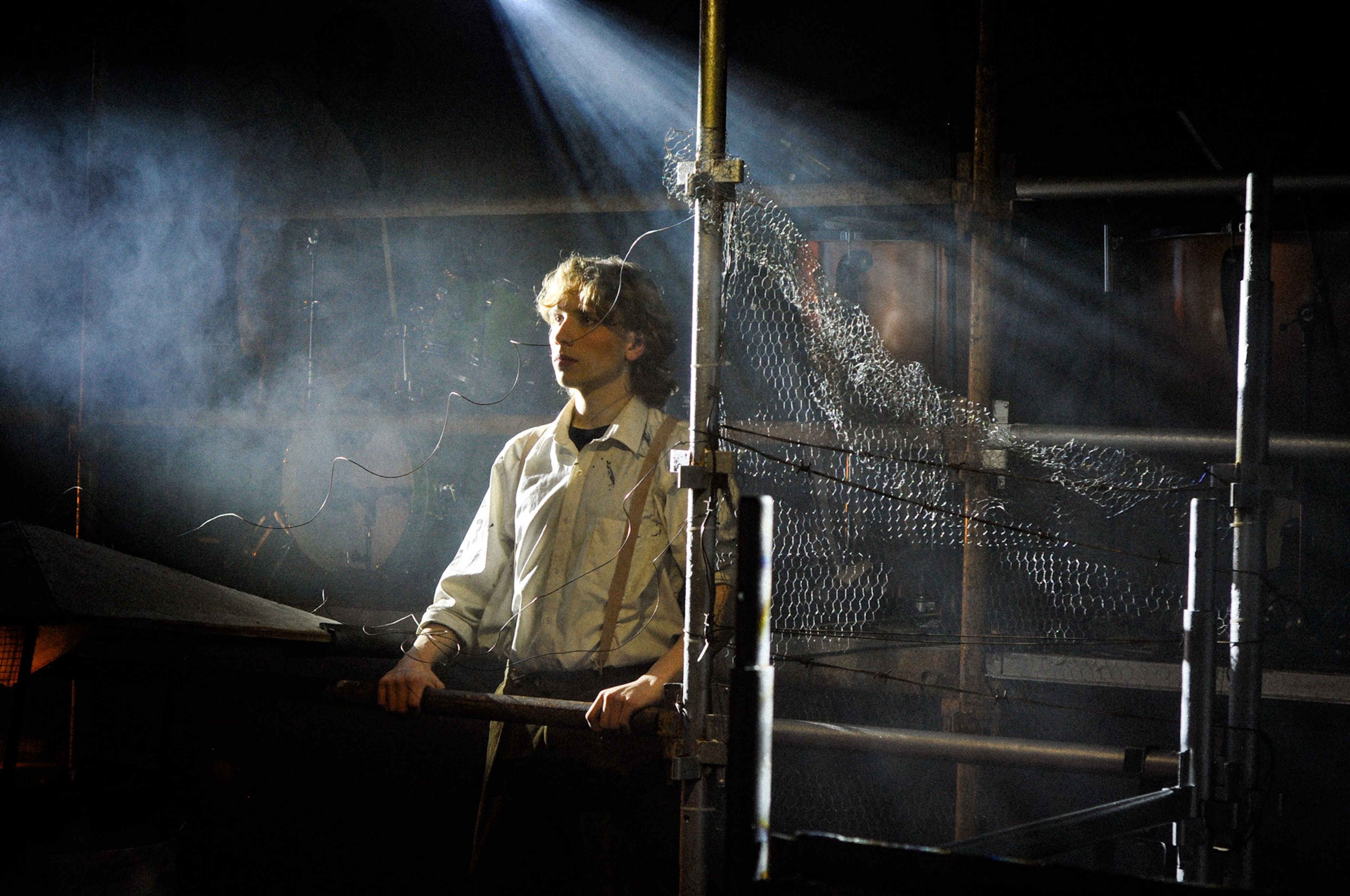 Theatre production photograph of a character standing on a scaffolding structure under a dramatic spotlight, with metal and wire mesh elements surrounding him in a smoky atmosphere.