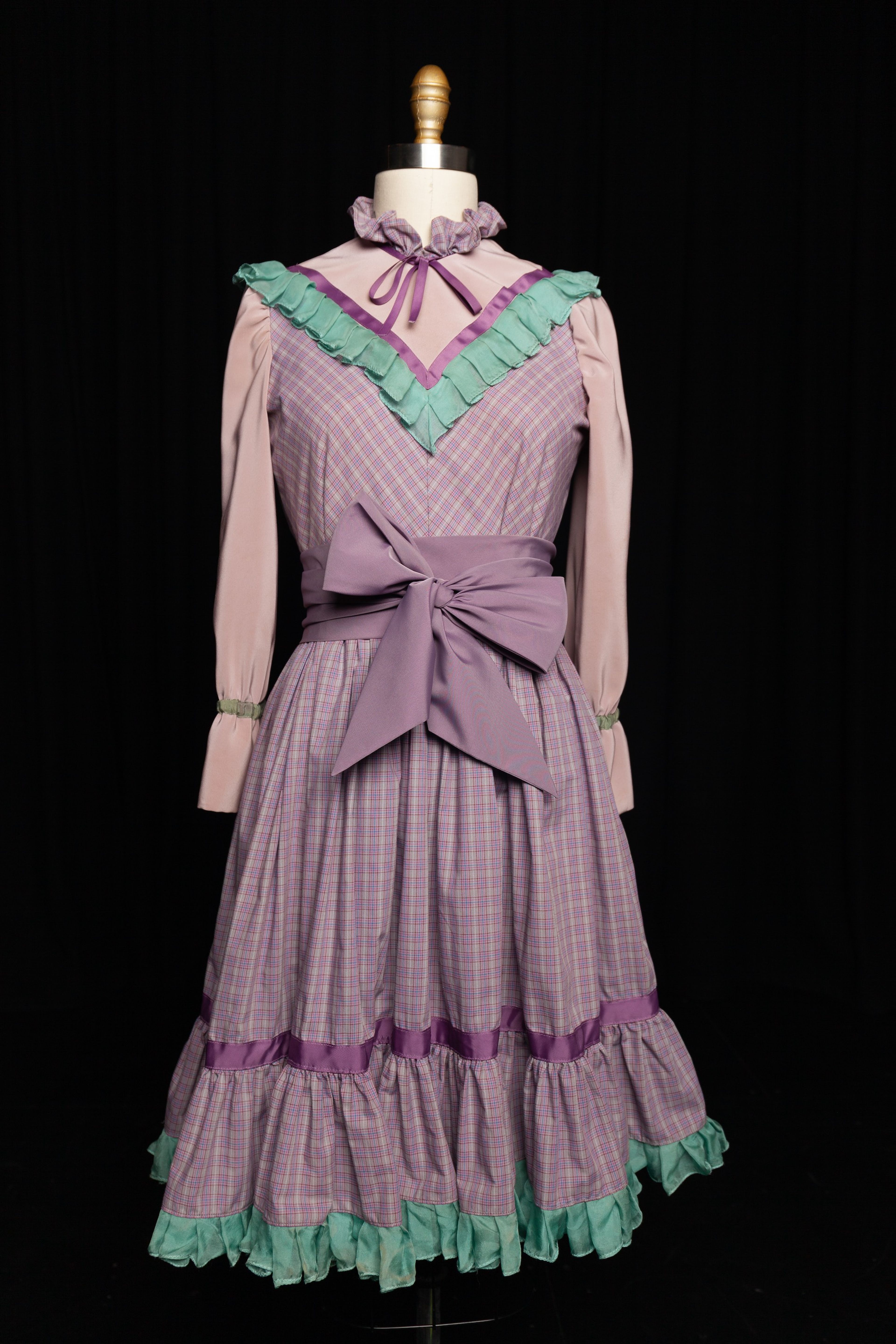 Costume photograph on mannequin showing a Victorian-style dress in lilac plaid with mint green ruffles, featuring a high ruffled collar, puffed sleeves, and large purple sash bow at waist.