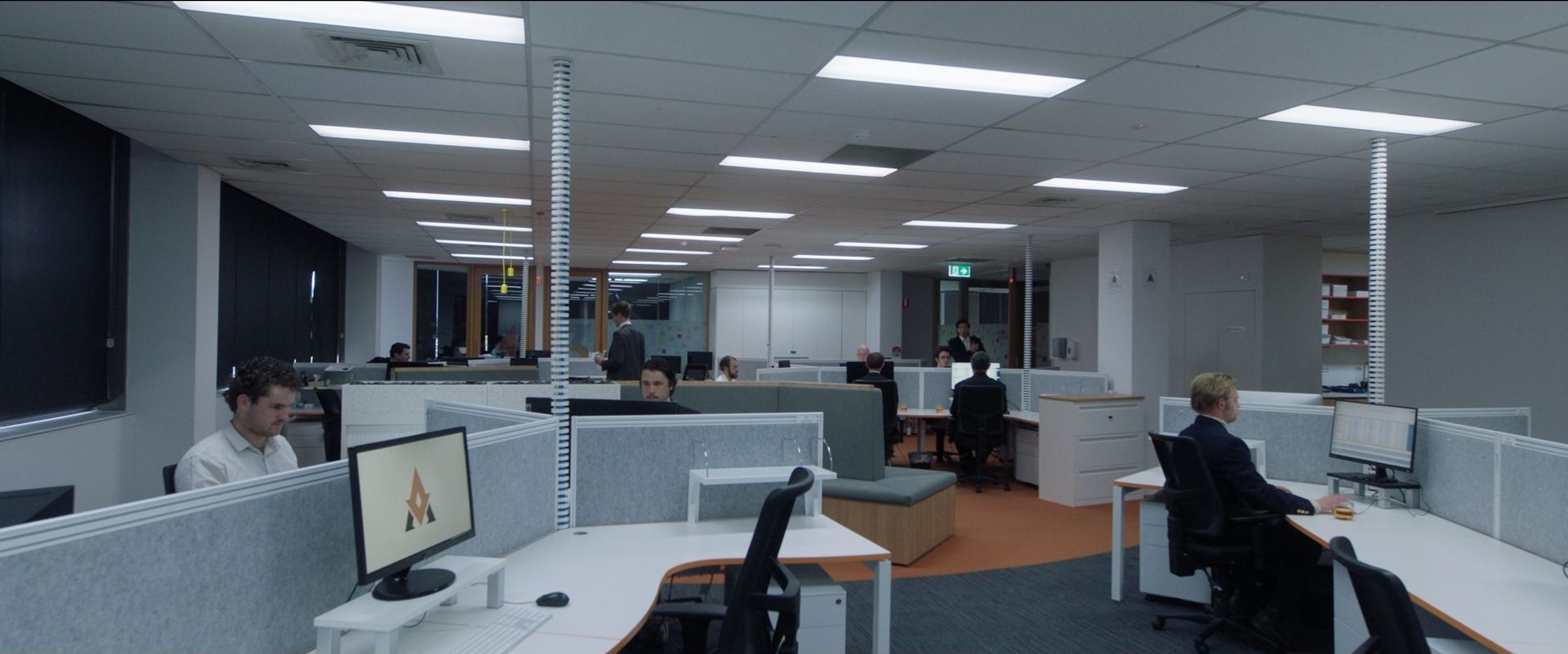 Film still of an open-plan office setting with workers seated at individual cubicles, typing on computers.