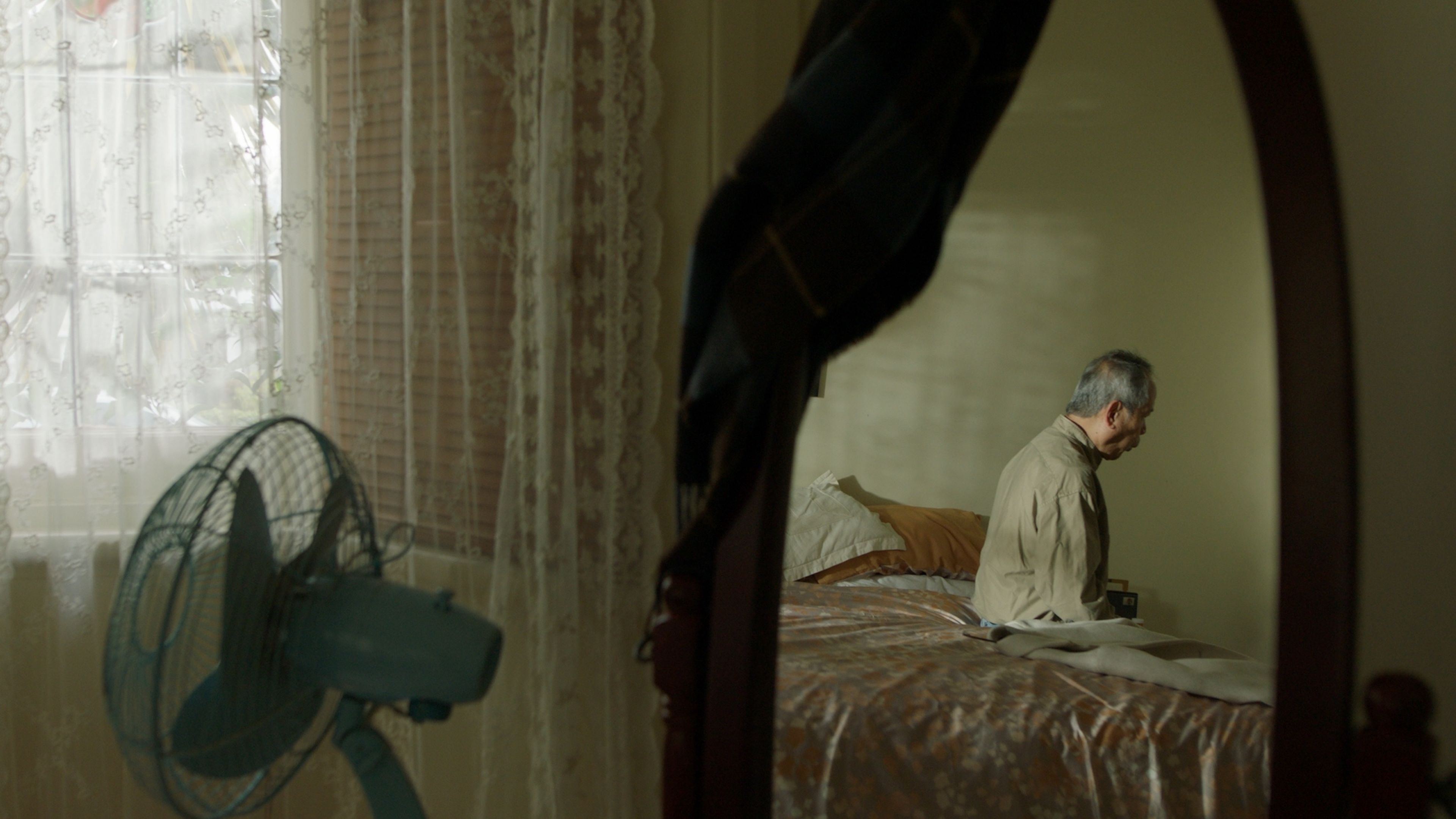 Film still of an elderly man sitting on a bed viewed through a doorway, with a vintage electric fan in the foreground and lace curtains filtering natural light