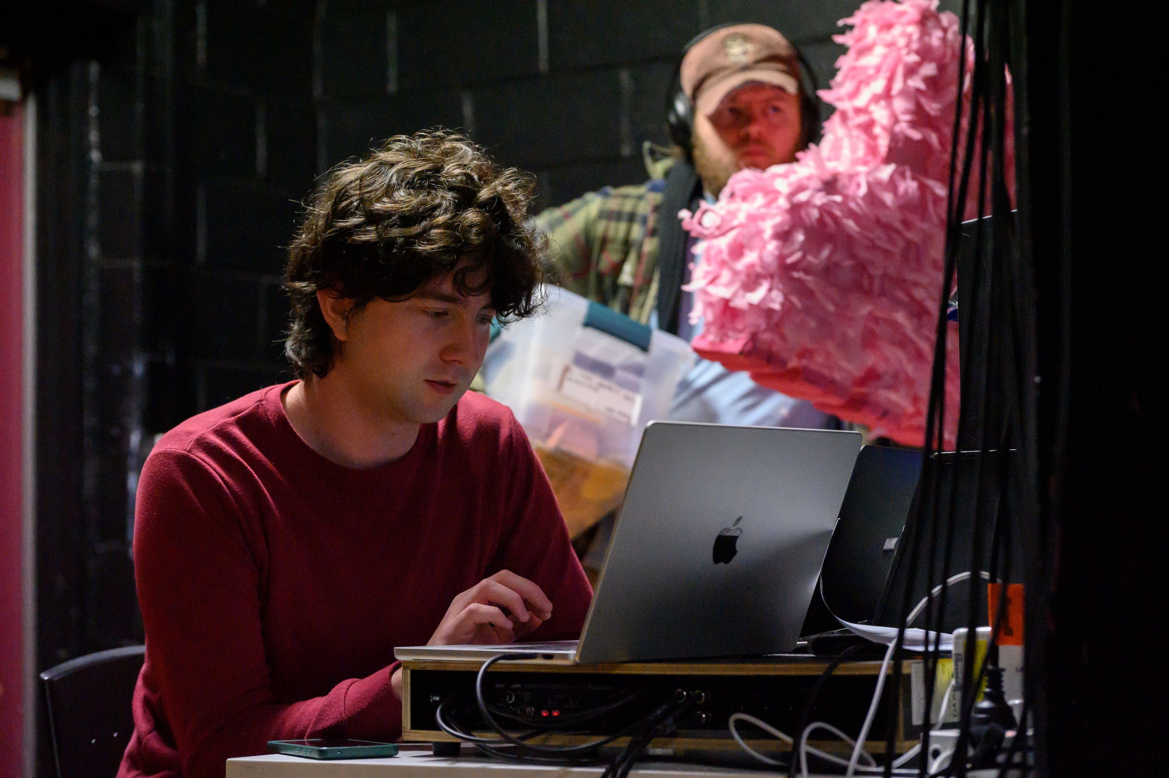 A person sits at a laptop backstage, focused on the screen, with another person in the background holding a pink costume prop.