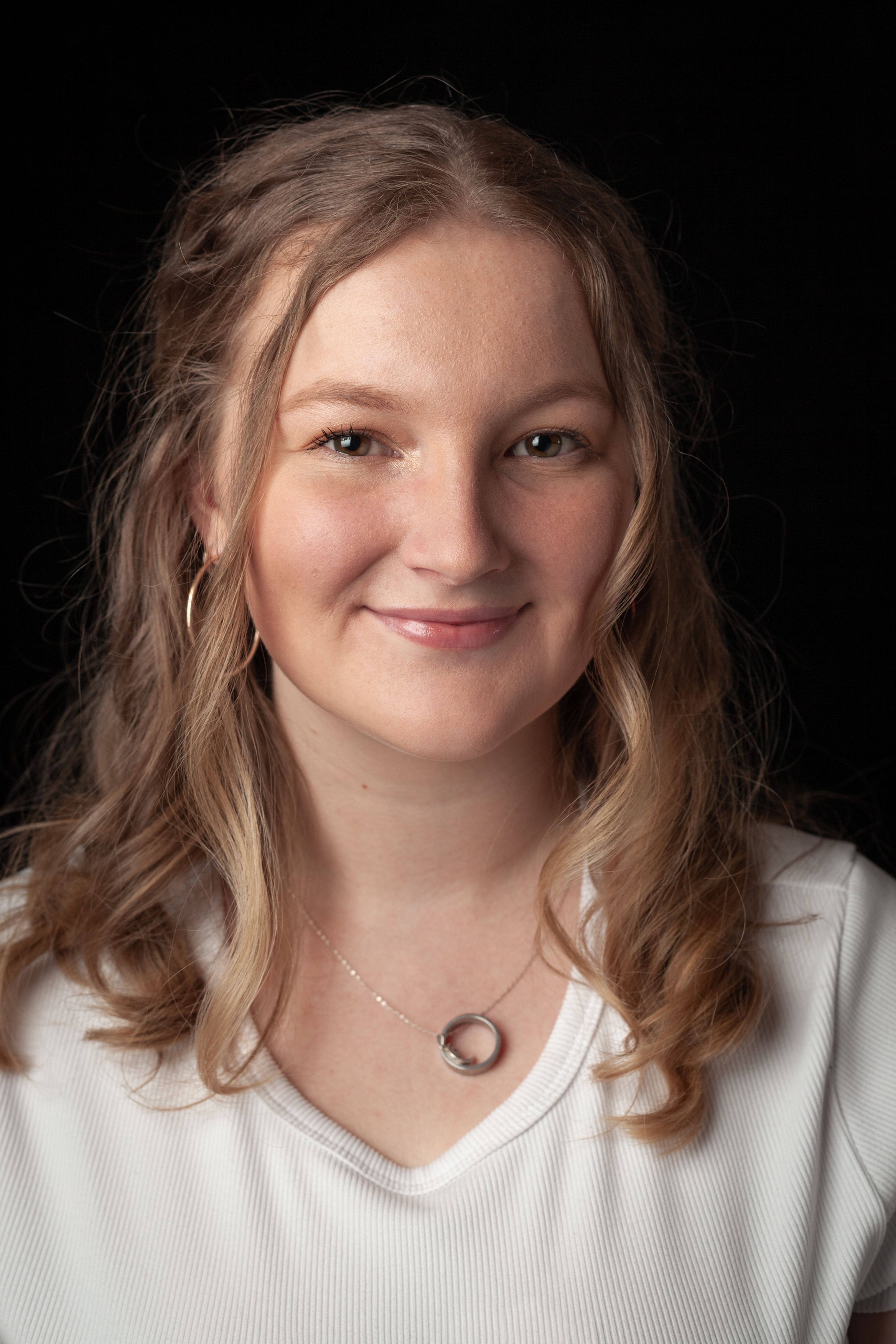 Headshot of a smiling young woman with wavy light brown hair, wearing a white top and a silver necklace, against a black background.