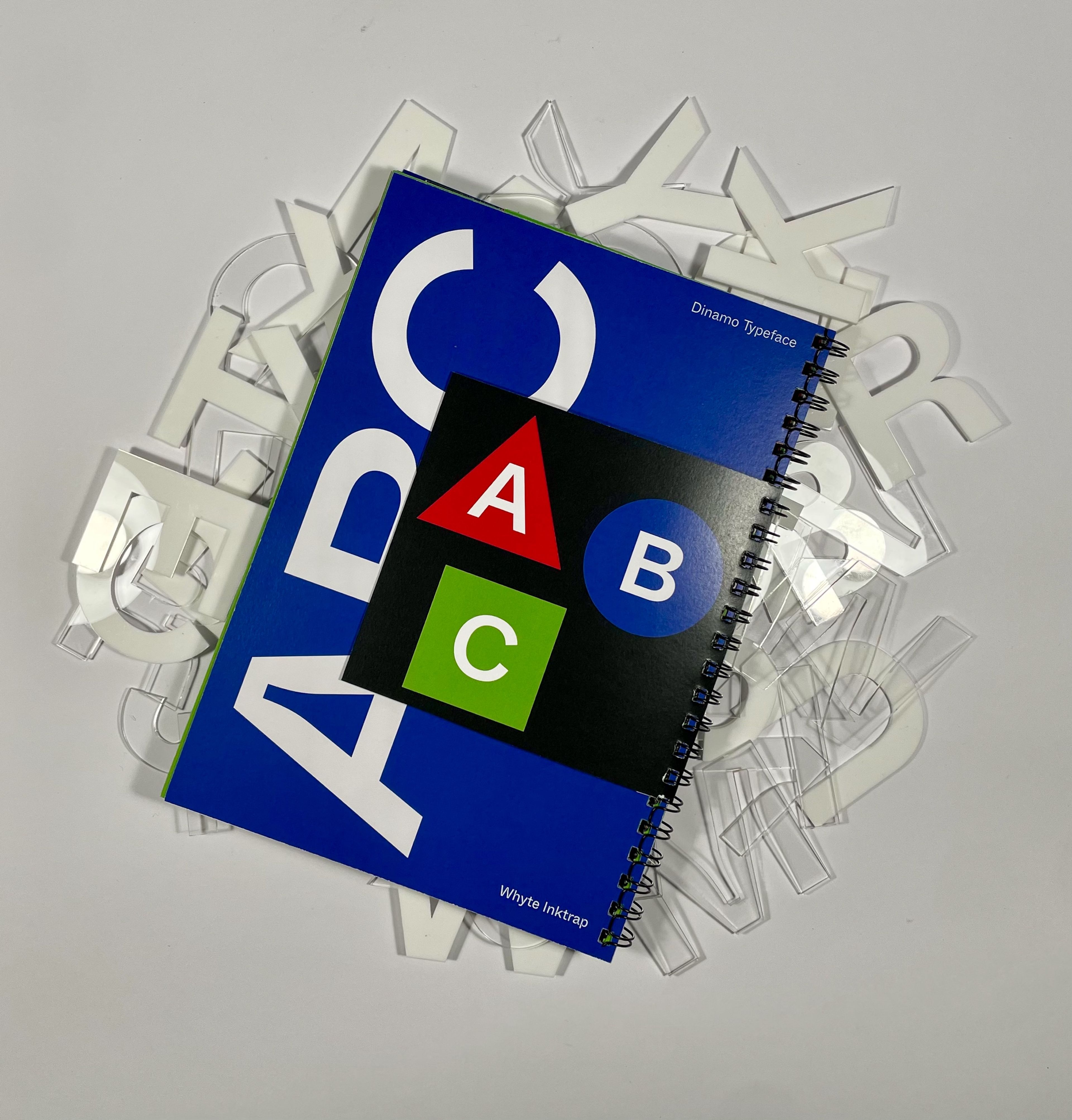 Cover of a typeface sample book titled 'Whyte Inktrap' by Dinamo, with a bold, blue cover displaying large white letters and an 'ABC' graphic in primary colours on a black square. Transparent acrylic letters are scattered beneath the book, adding a playful touch.