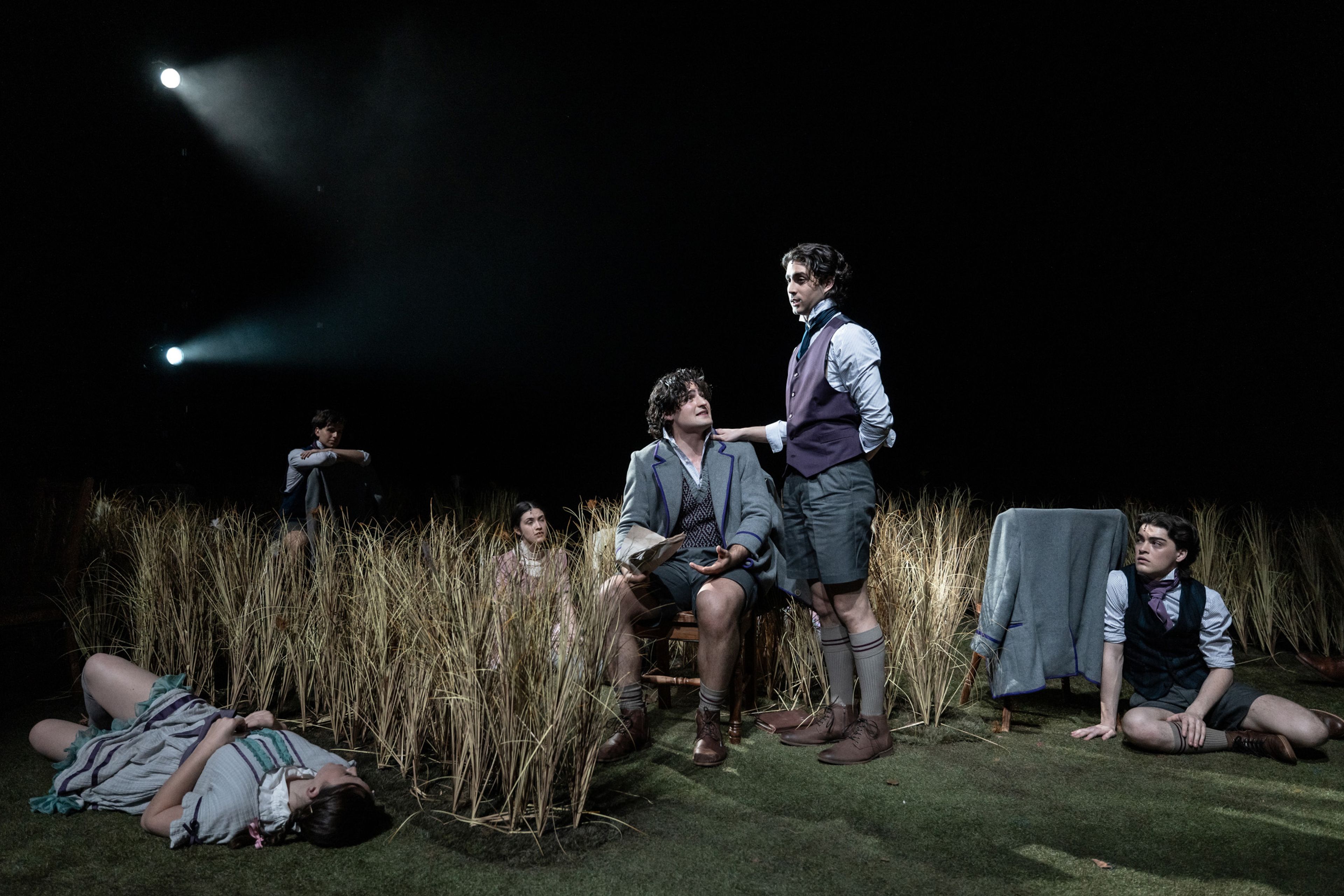 Theatre production photograph showing schoolboys in period uniforms lounging among wheat grass, with two performers in conversation centre stage and dramatic spotlighting above.