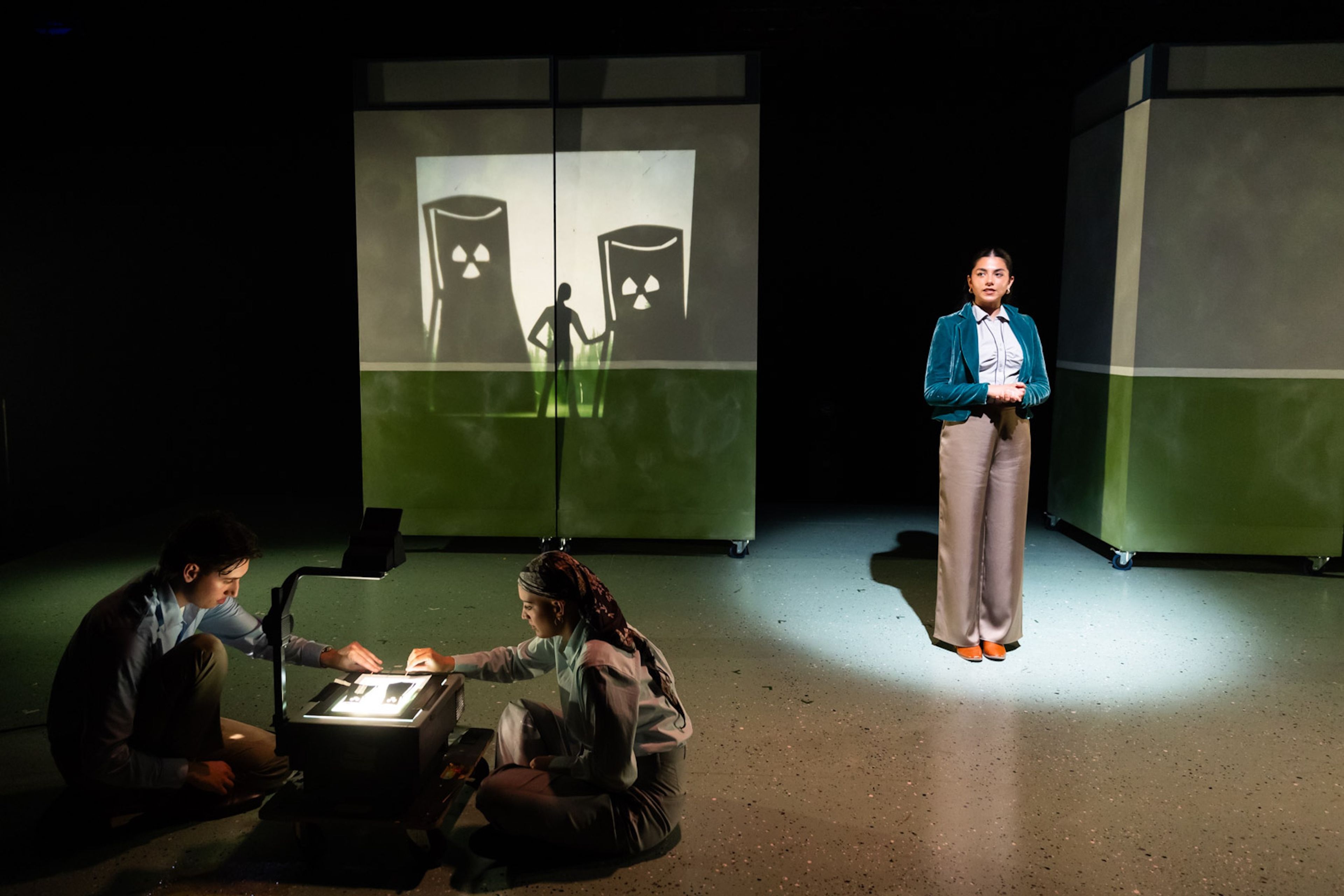 Theatre production photograph with one performer in spotlight wearing a teal blazer, standing beside projected images of radioactive symbols, while two other performers work with an overhead projector on stage.