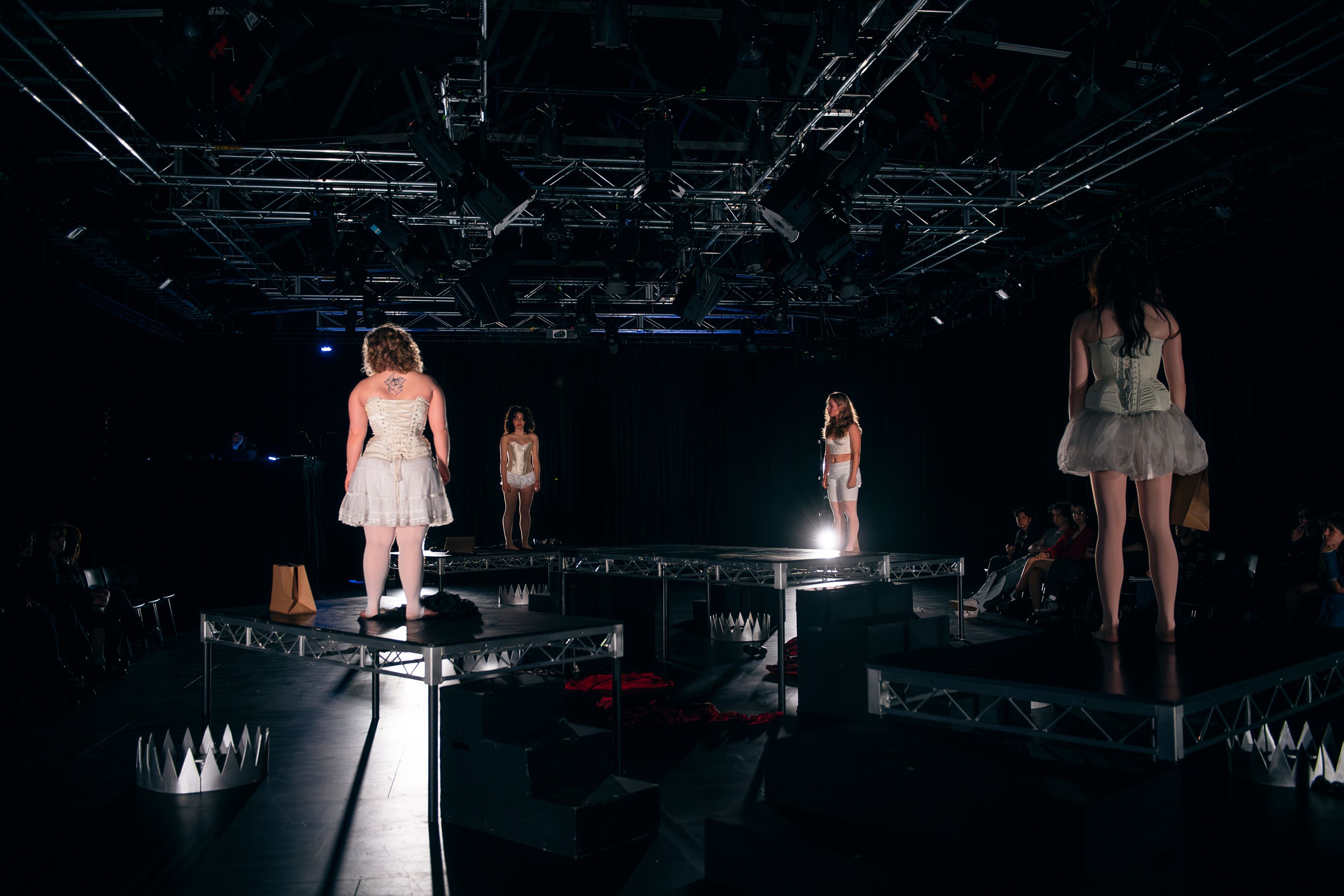 Theatre production photograph with actors in corset costumes positioned on raised platforms across the stage, each in their own spotlight, with a dark, dramatic atmosphere.