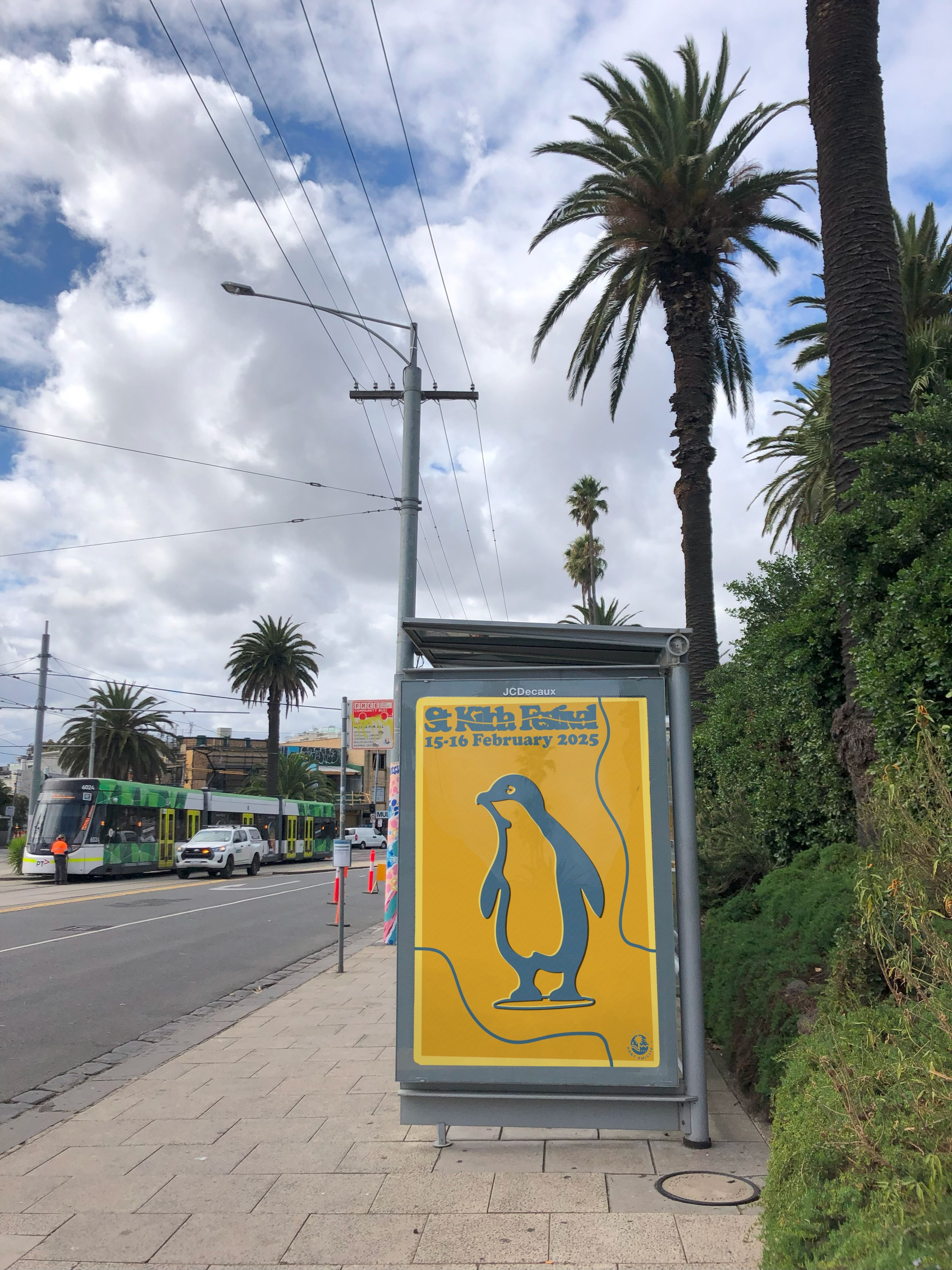 Yellow and blue "St Kilda Festival" poster with penguin illustration and February 15-16, 2025 dates, set at a tram stop with palm trees and passing tram.