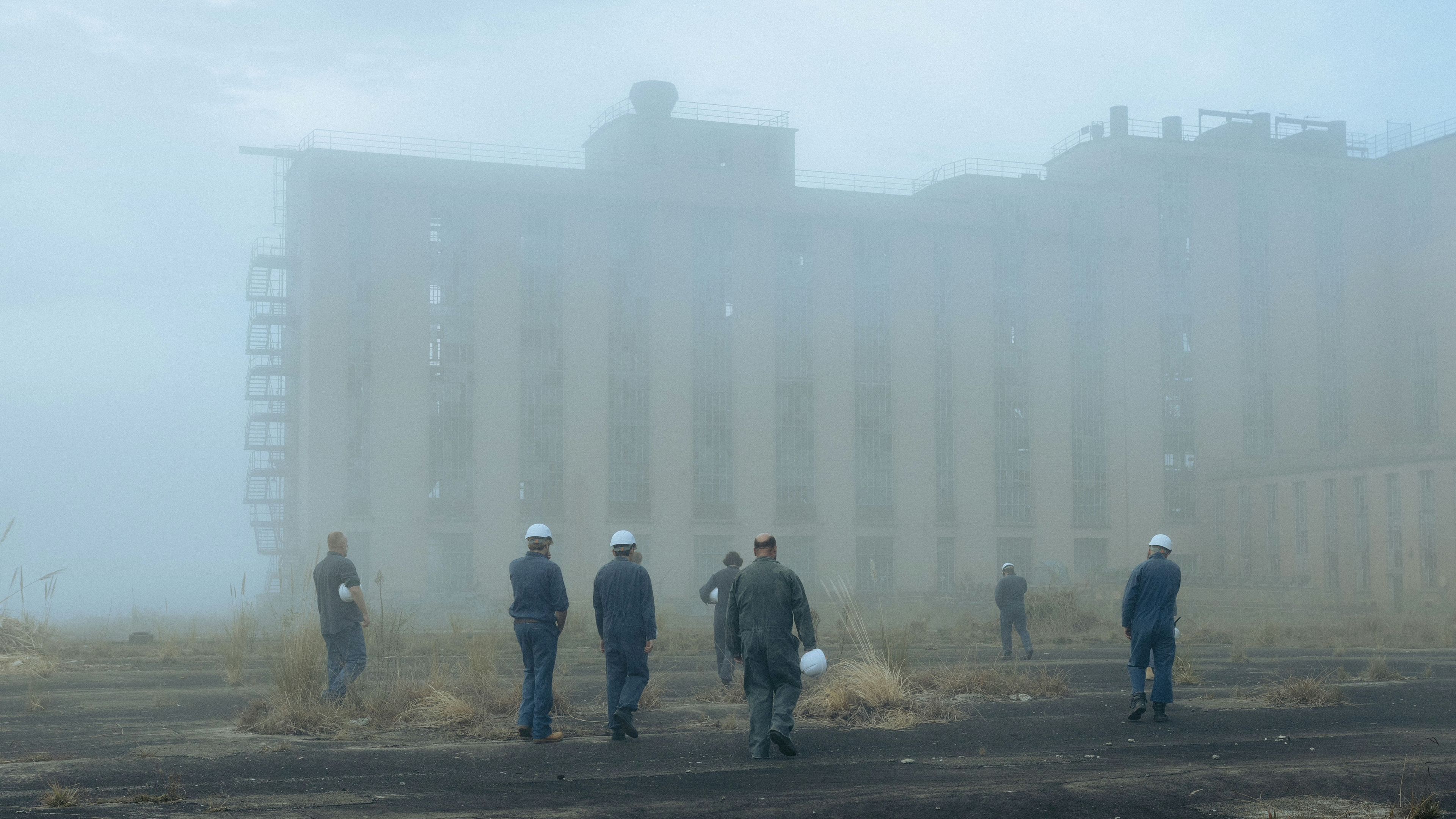 Film still showing workers in hard hats walking towards a large industrial building shrouded in fog, creating an eerie atmosphere.