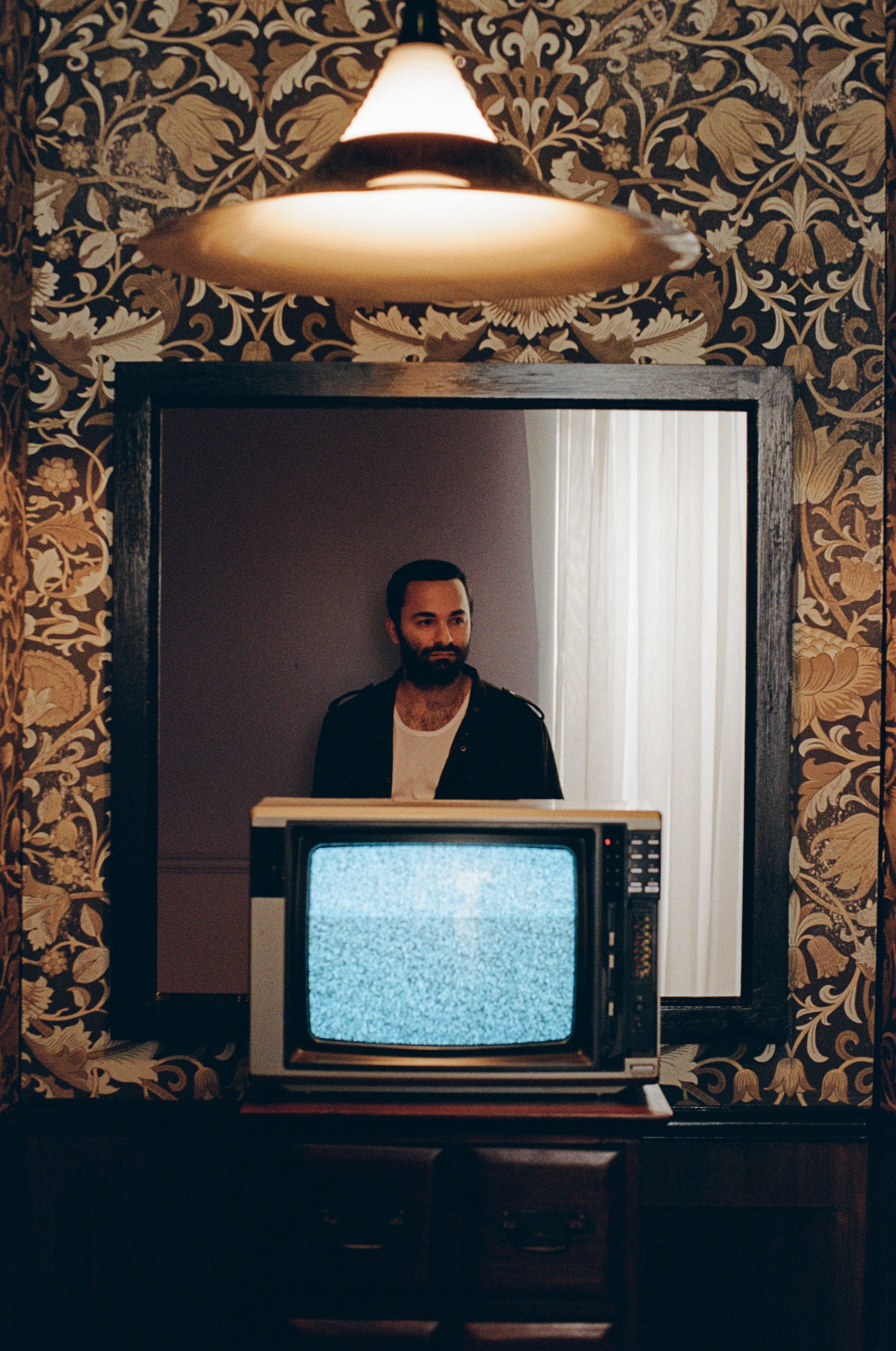 A retro television set with static on screen, framed by ornate wallpaper and pendant light, with a figure visible in a mirror behind.