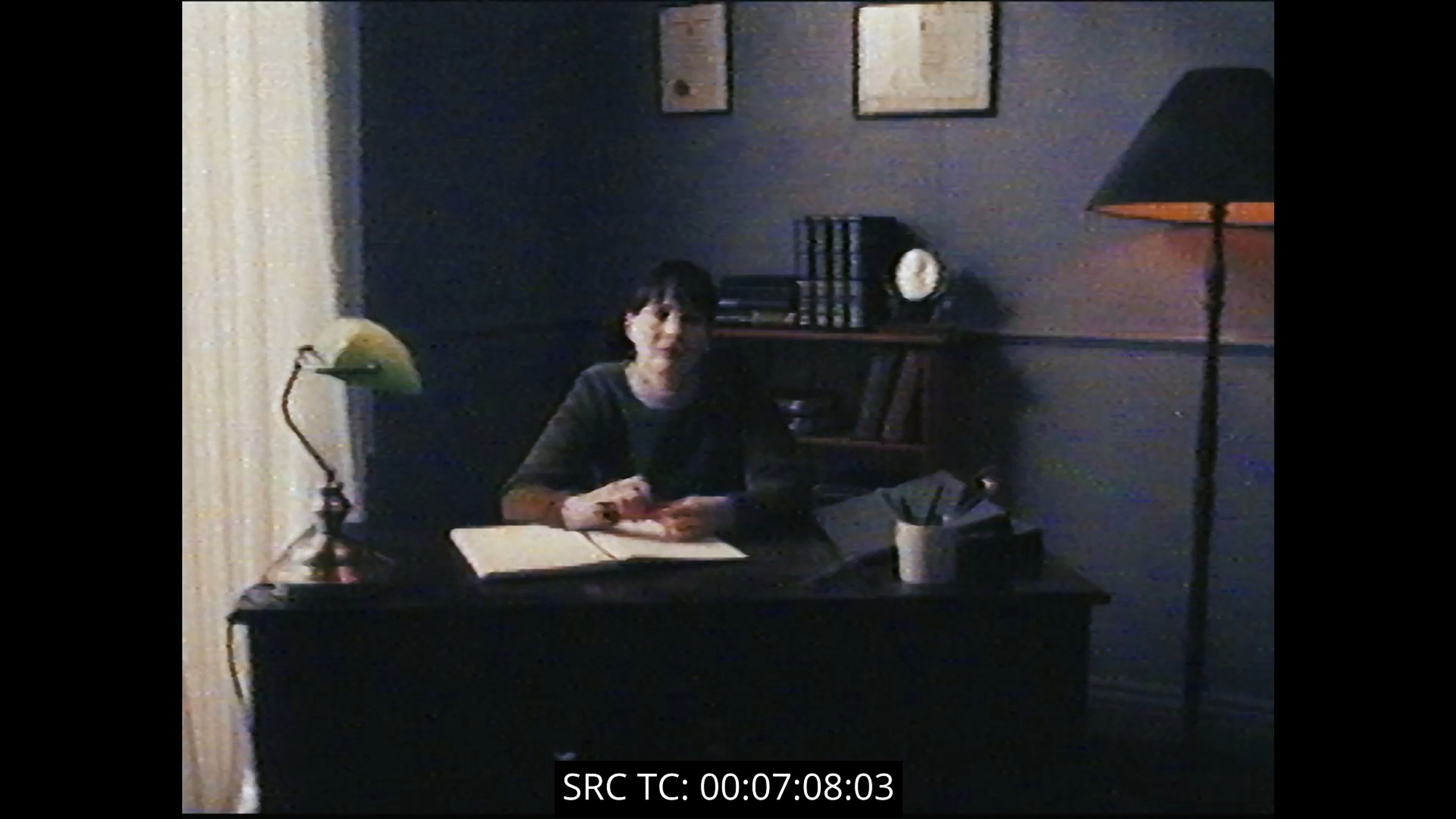 Film still shot on vintage video showing a woman at a wooden desk with banker's lamp, framed by bookshelves and wall art, with timecode overlay.