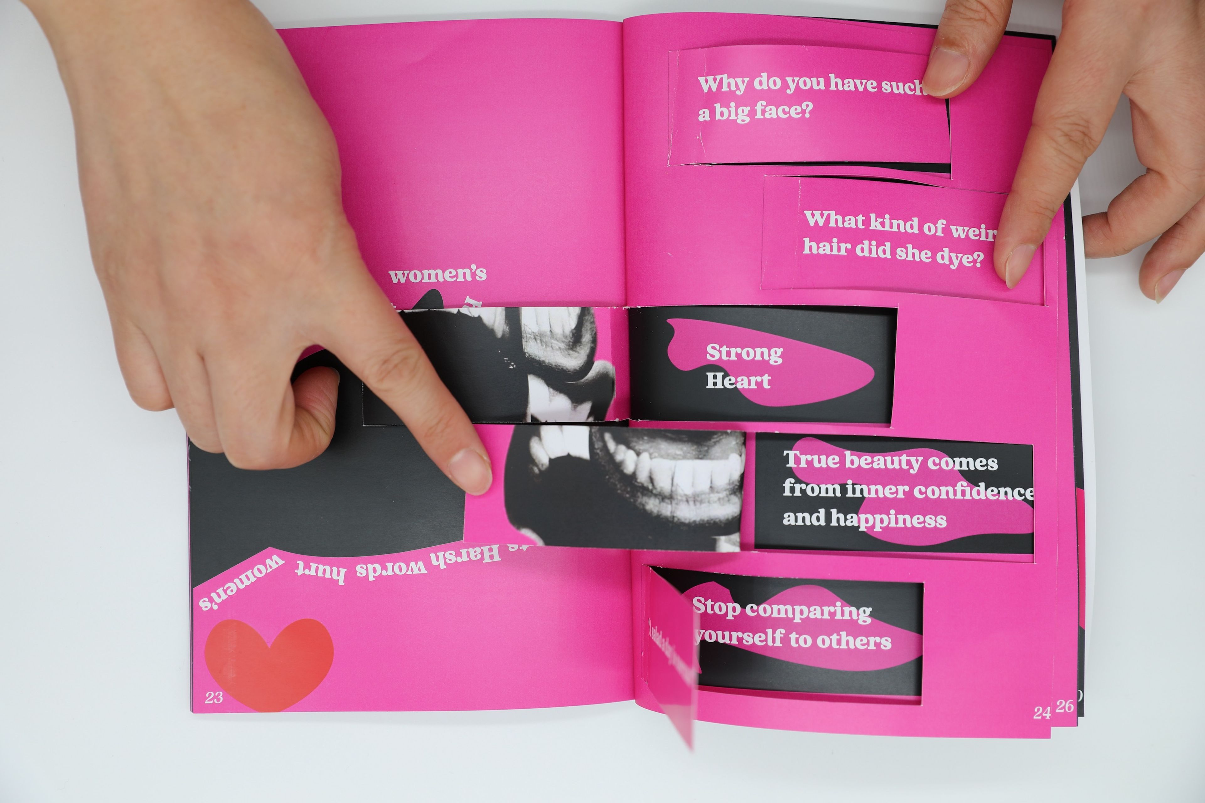 Bright pink and black spread from an art book, with interactive flaps that reveal affirmations such as 'Strong Heart' and 'True beauty comes from inner confidence and happiness.' Surrounding text questions stereotypical beauty standards with statements like 'Why do you have such a big face?'