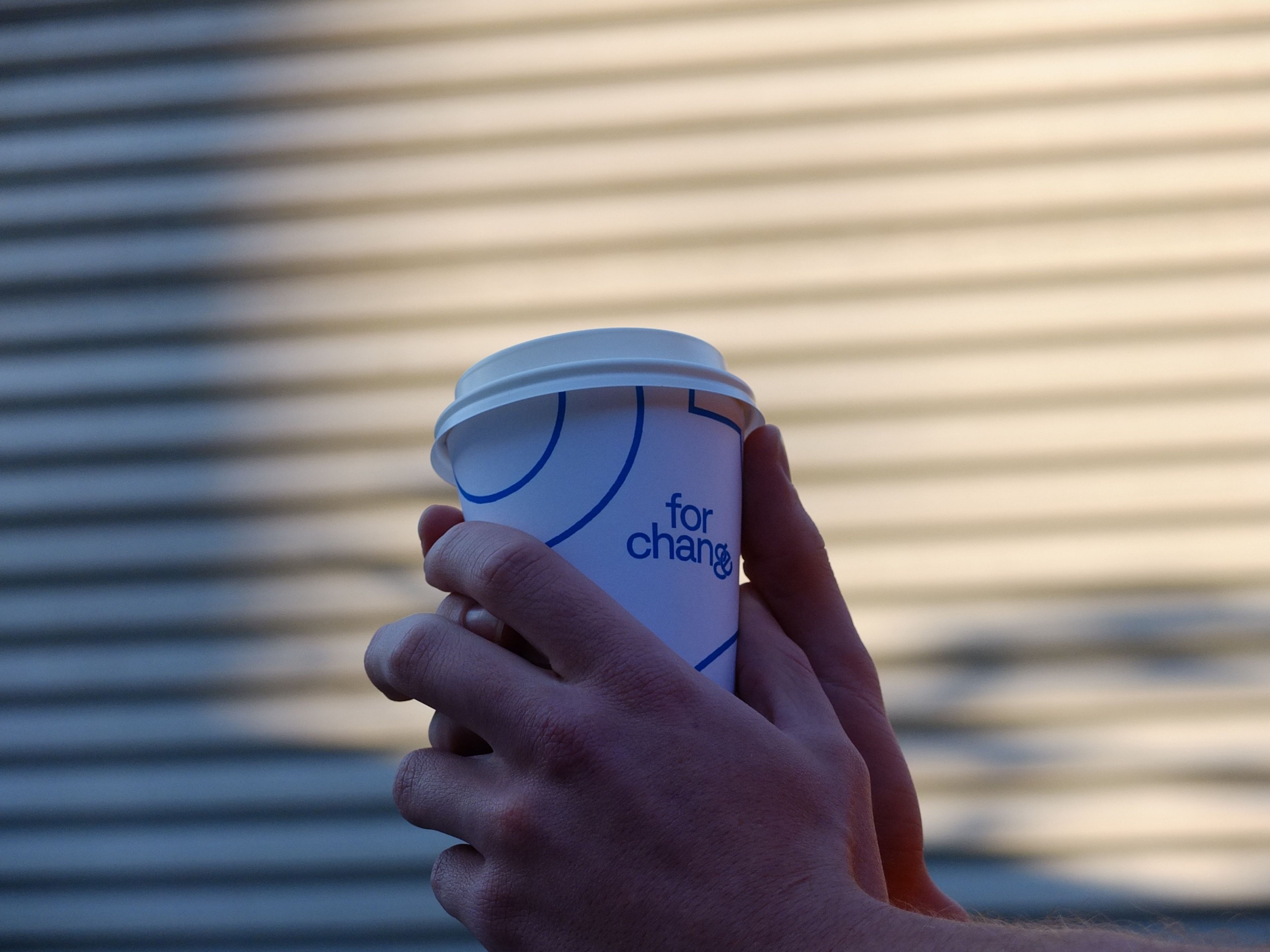 A close-up of hands holding a paper coffee cup against a softly blurred background. The cup features the words "for change" in blue on a white background, suggesting a minimalist, socially-conscious brand design.