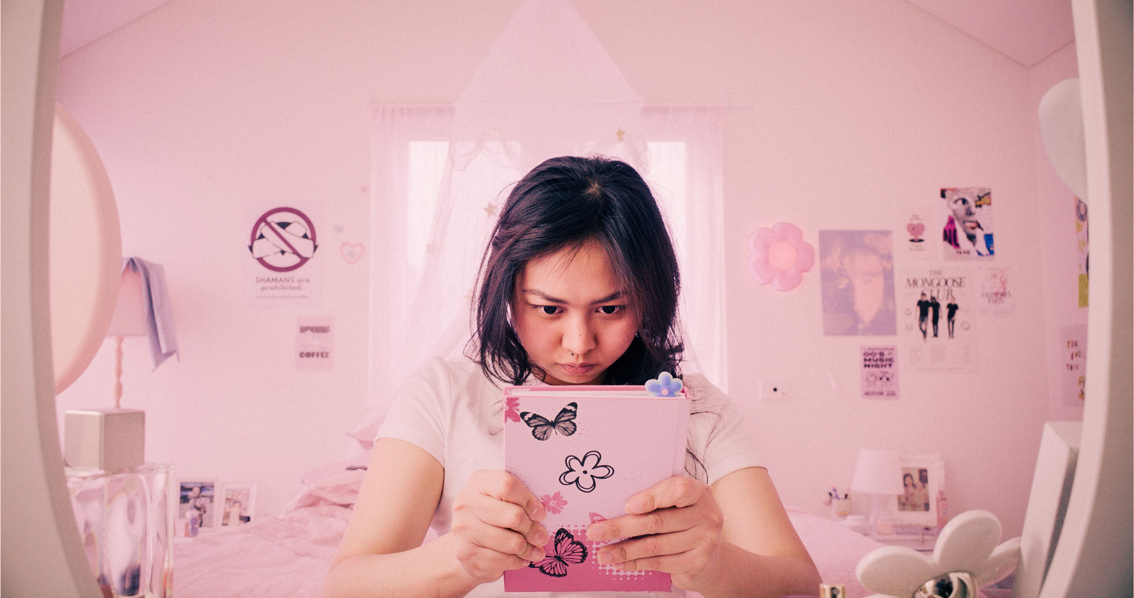 A young woman in holding a pink diary decorated with butterflies and flowers. She sits in a pastel pink bedroom adorned with soft lighting, floral and poster decorations