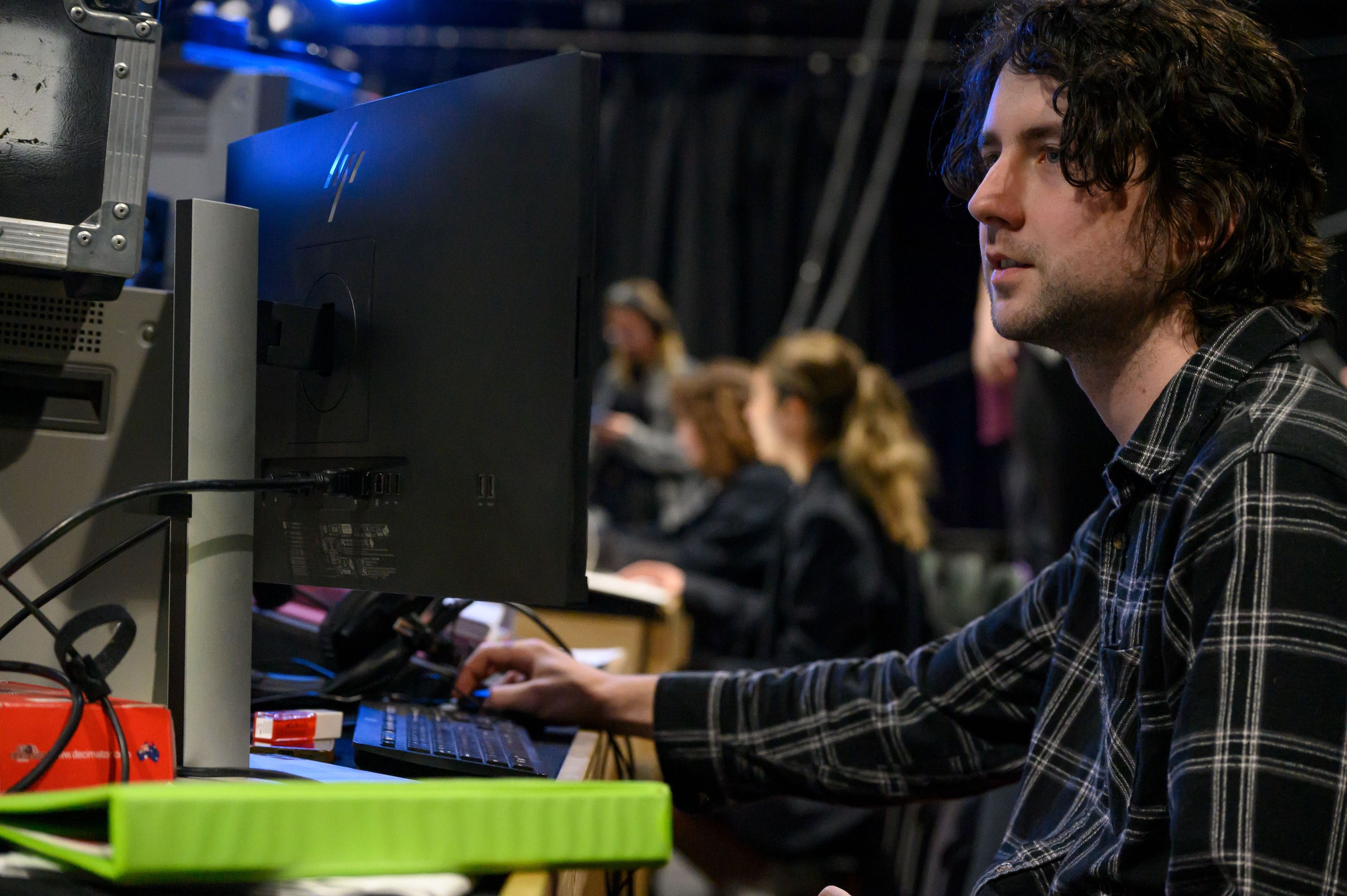 A person in a plaid shirt works at a computer in a backstage setting, with others visible in the background.