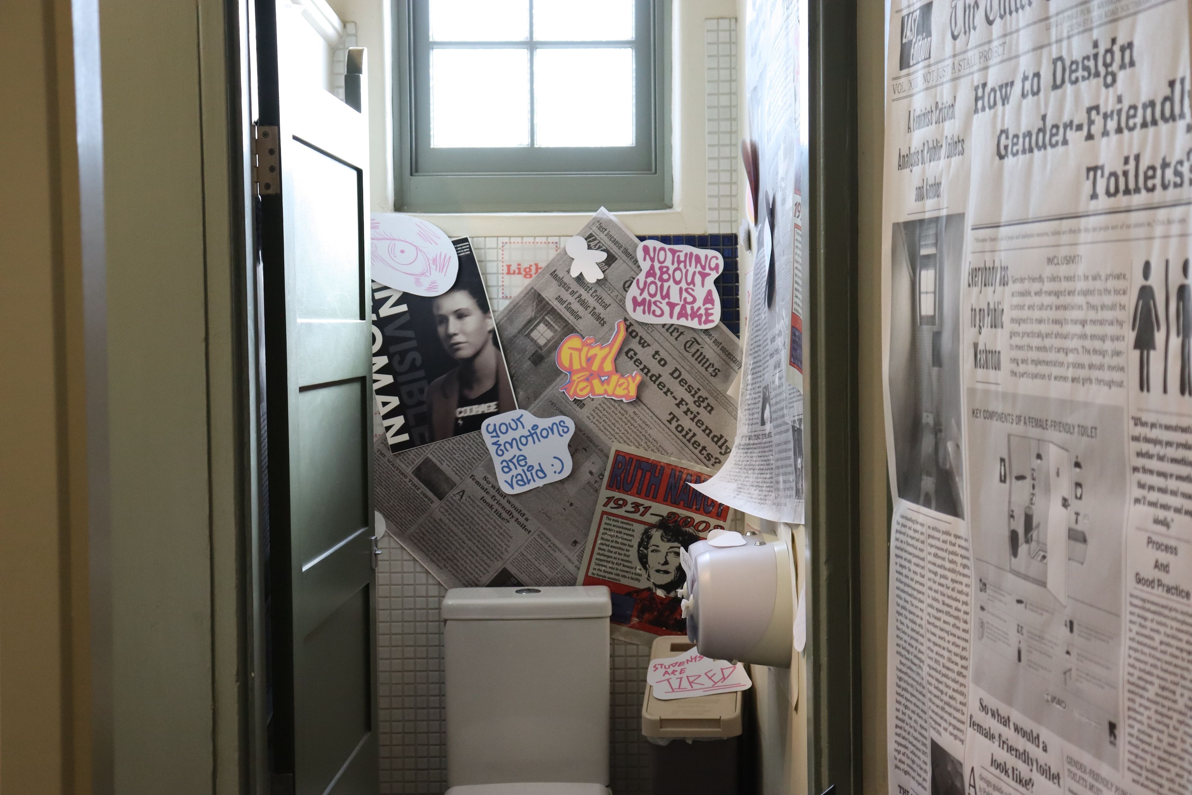 Restroom interior with sage green door, with walls adorned with collaged newspaper articles and handwritten affirmation stickers.