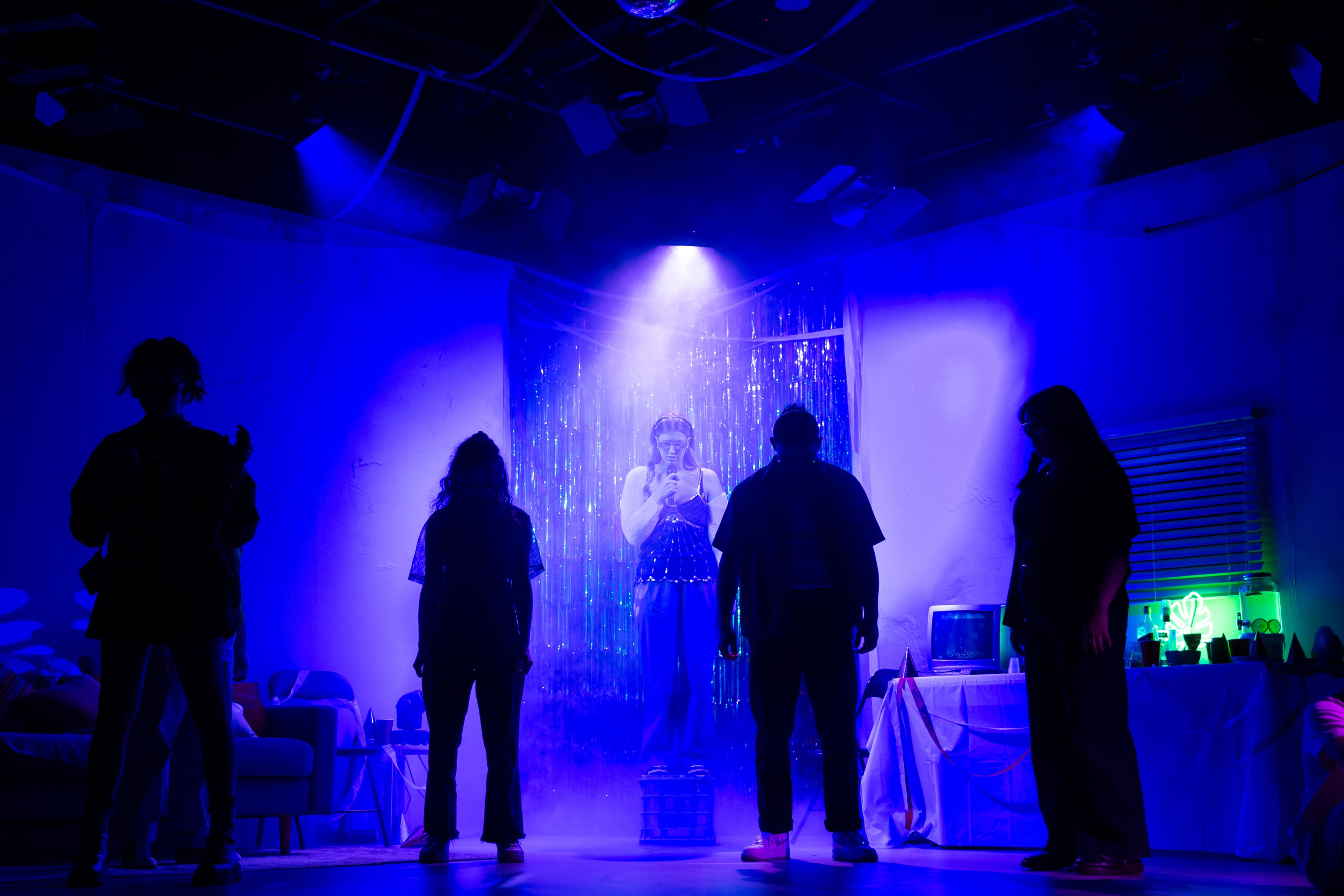 Theatre production photograph showing a performer in costume silhouetted against a silver tinsel backdrop, surrounded by other performers in black, with intense blue lighting creating atmosphere.