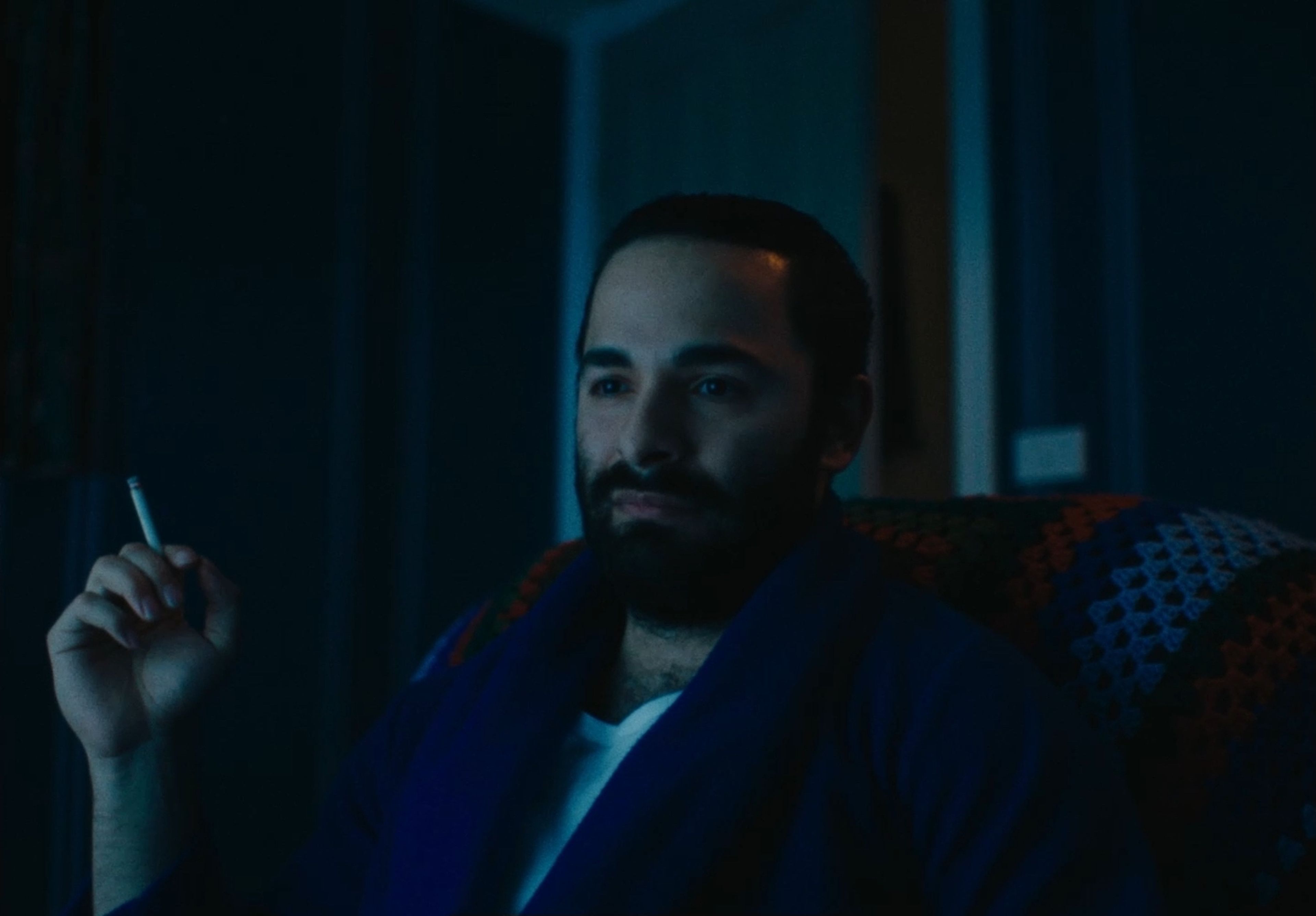 Film still portrait shot lit in deep blue tones, showing a bearded man in a navy cardigan holding a cigarette, against a darkened interior.