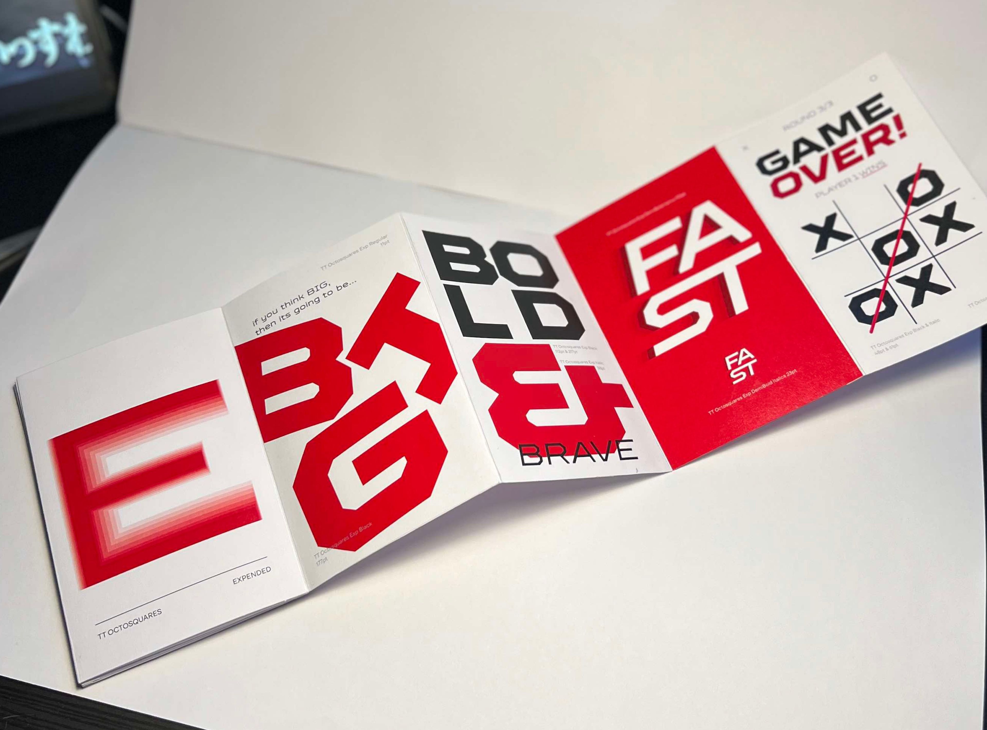 Open type specimen booklet showcasing large, bold typographic samples in red, black, and white. Words like 'BIG,' 'BOLD,' 'BRAVE,' and 'FAST' are displayed in dynamic layouts, with a 'GAME OVER!' page styled like a tic-tac-toe board, capturing an energetic and assertive theme.