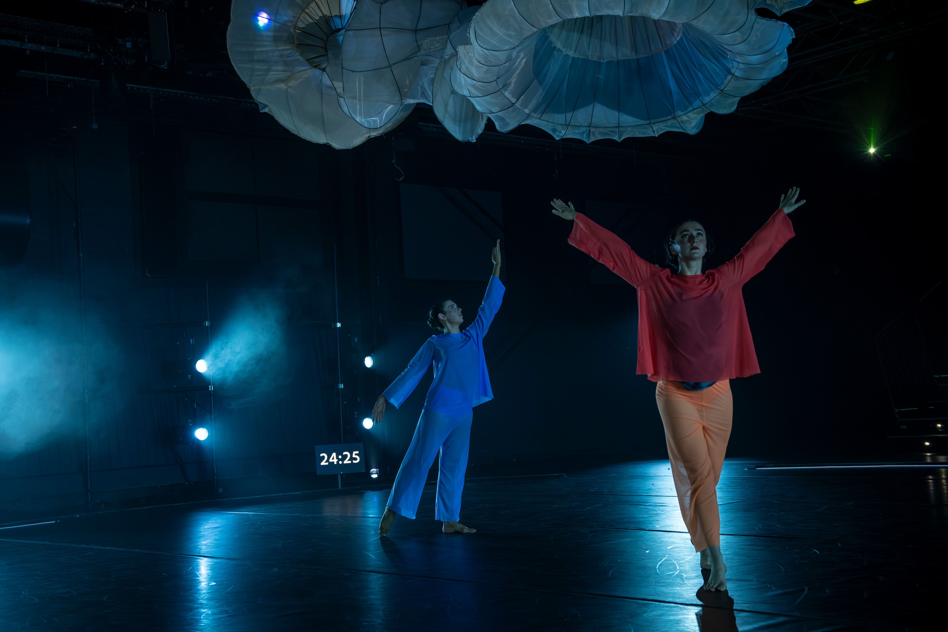 Theatre production photograph showing two dancers in contrasting costumes – one in blue and the other in orange – reaching upwards with a misty blue backdrop and large fabric structures overhead.