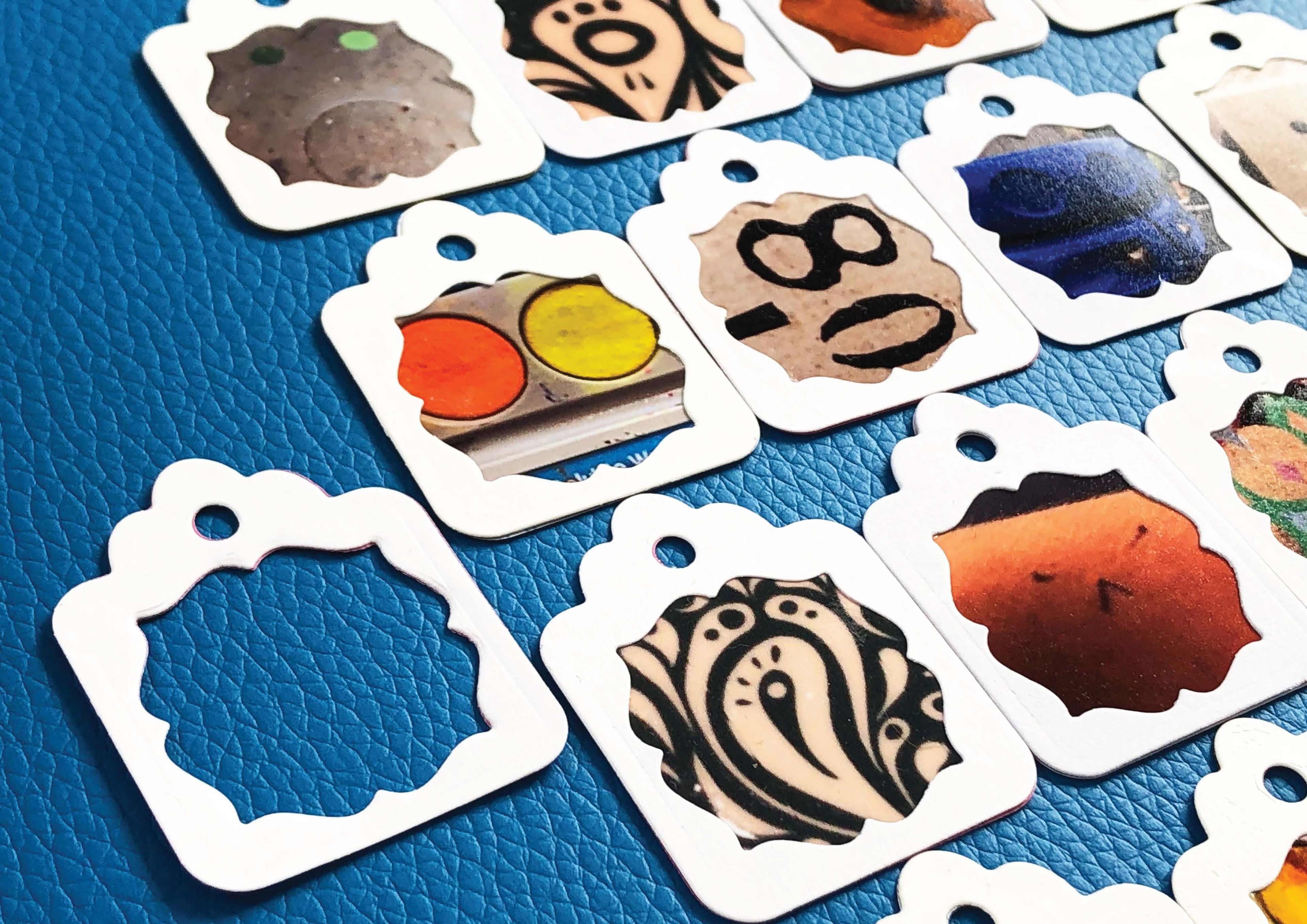 Collection of small paper tags on a blue textured background, each with a cut-out frame displaying close-up images. Designs within the frames include vibrant paint swatches, abstract patterns, and organic shapes.