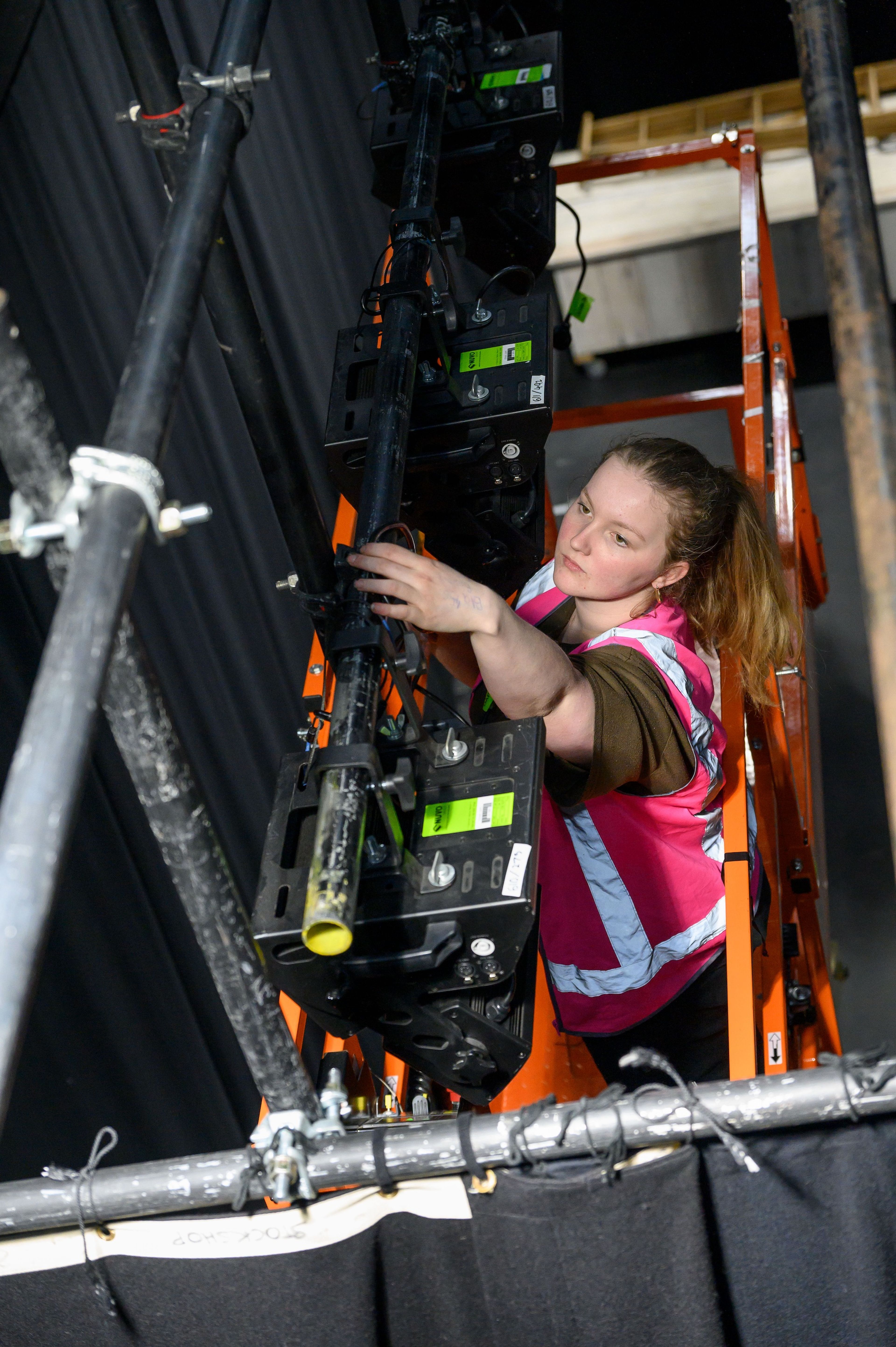 Crew member in a high-visibility vest adjusting stage lights while standing on an orange ladder, focused on setting up equipment above the stage.