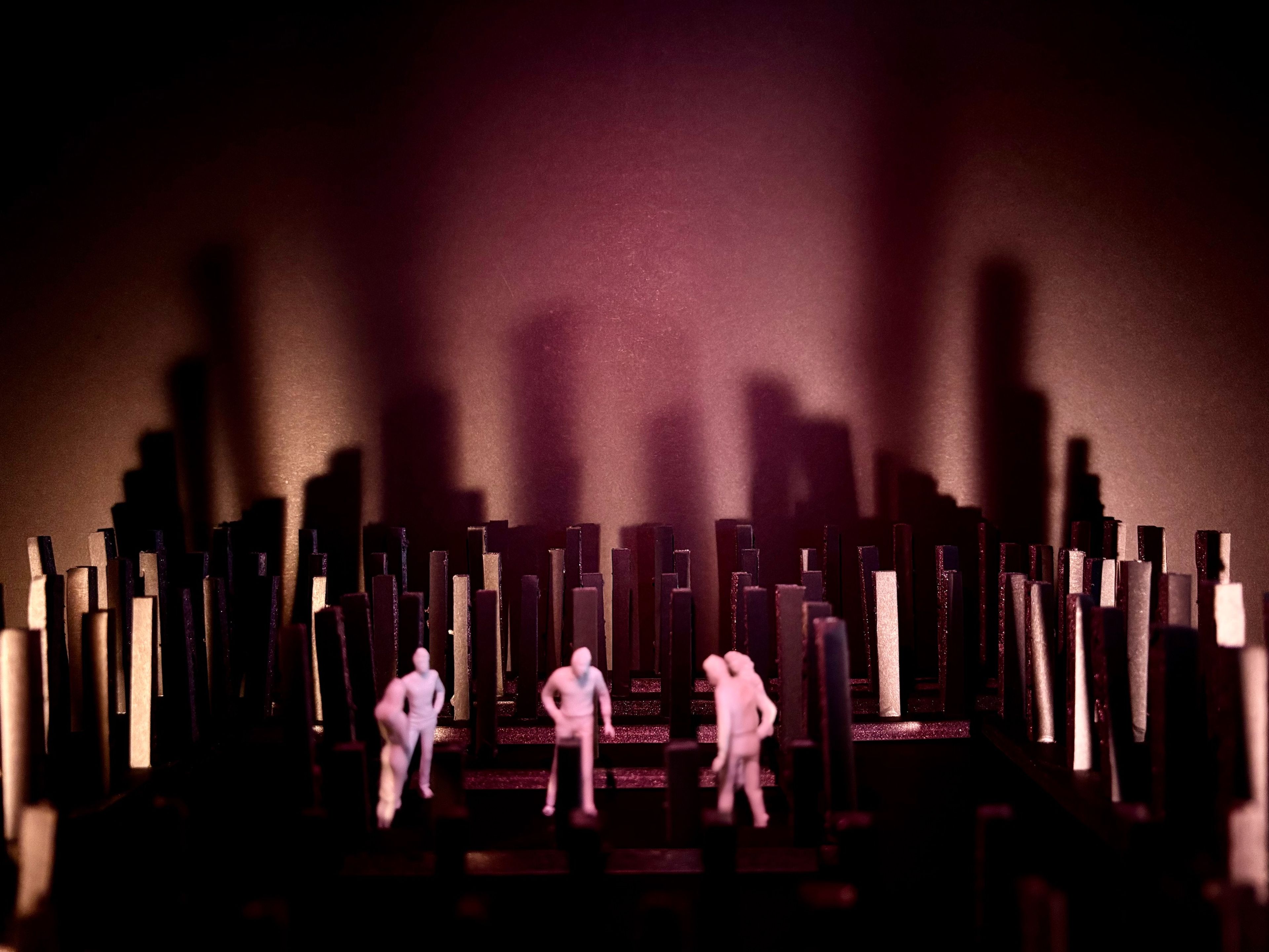 Set model with three figures in a dense field of vertical black structures, lit by a soft pink spotlight.