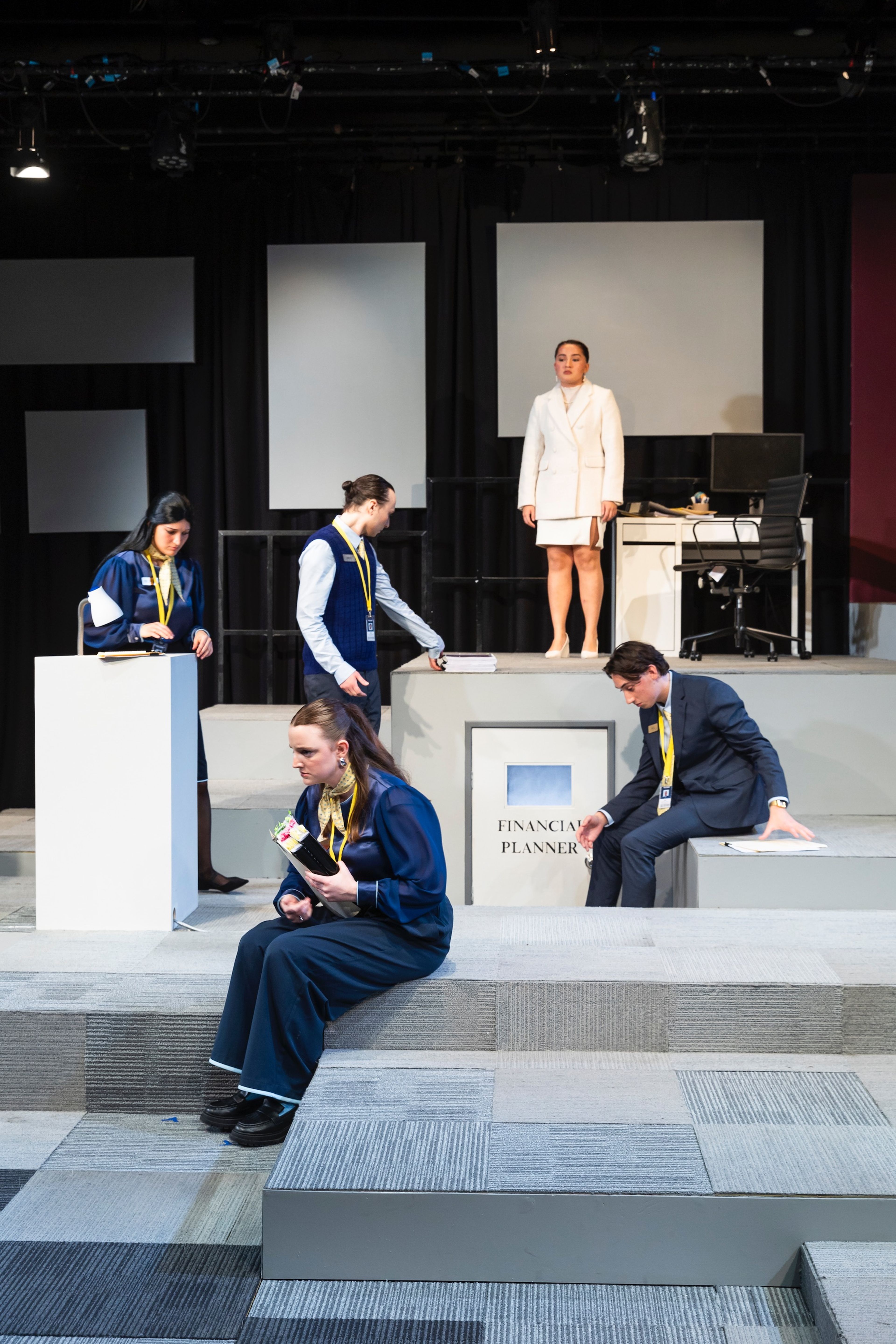 Theatre production photograph showing actors in a corporate setting, with one character standing on a platform in a white suit while others interact with office props.