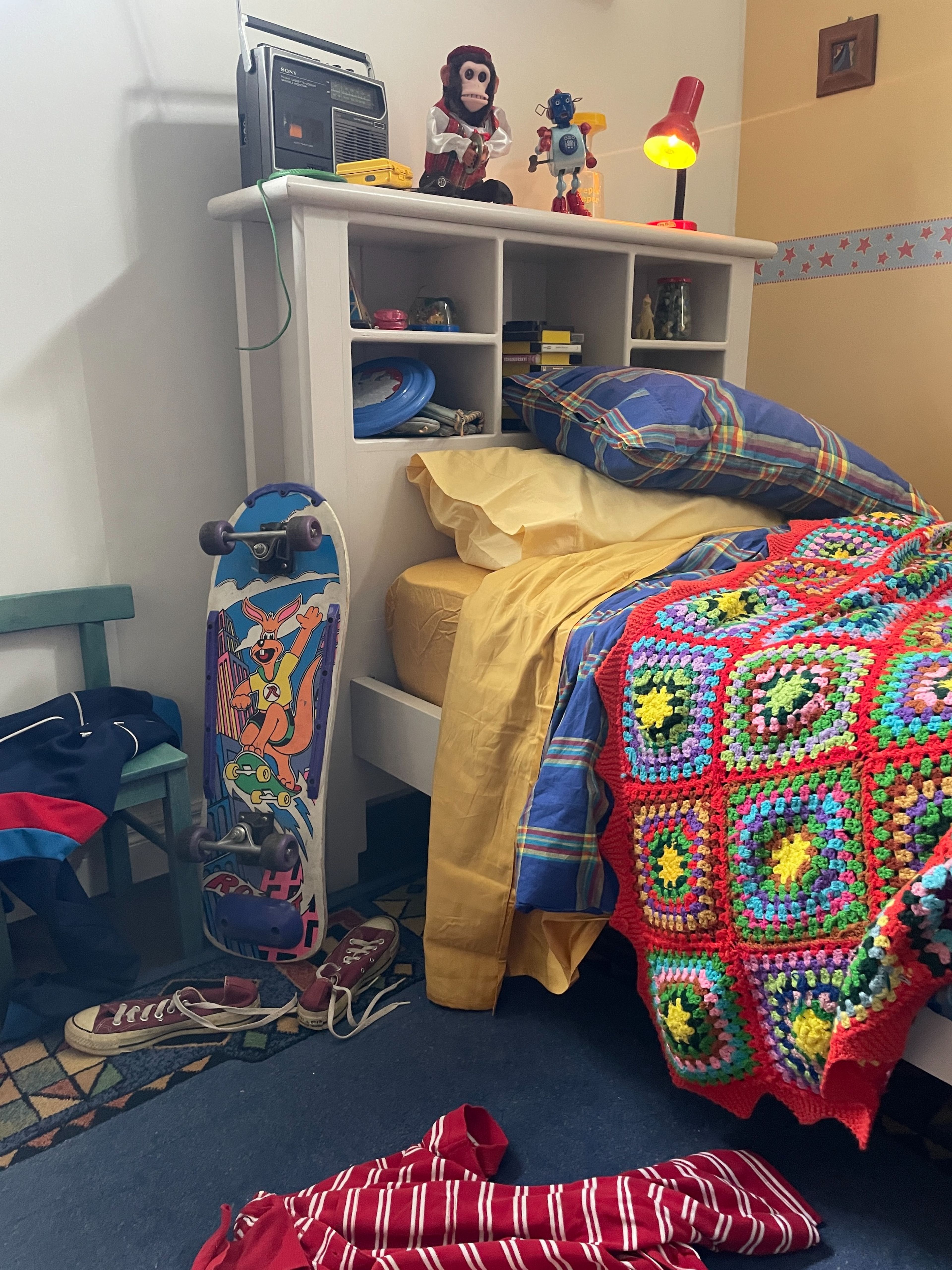 Set dressing photograph of a 1980s teenager's bedroom, featuring a skateboard, vintage radio, toys on a white bookshelf headboard, and a colourful crocheted blanket on the bed.