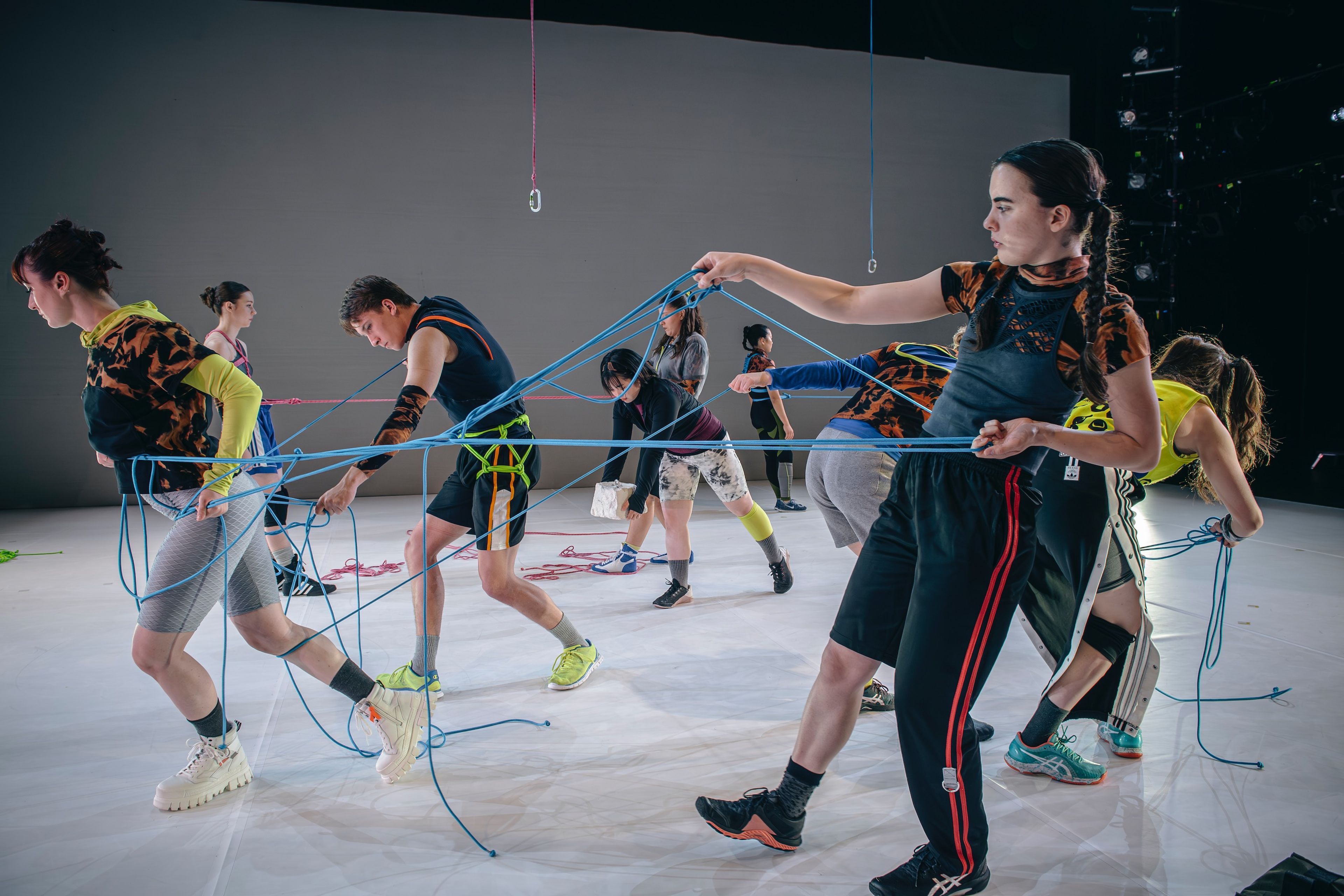 Dancers in athletic wear connected by a web of blue and pink ropes, on a white floor with hanging ropes visible above.