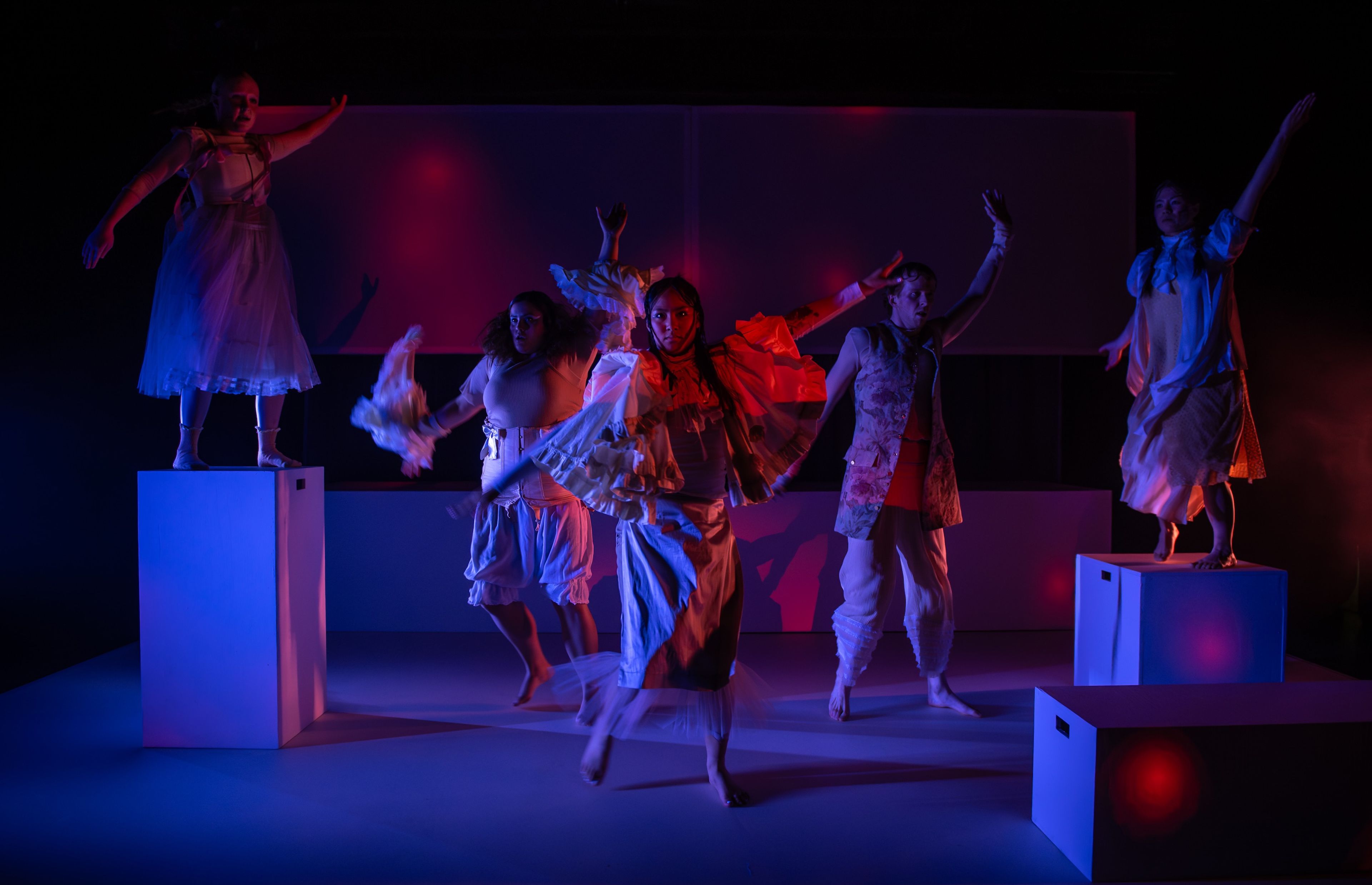 Performance photograph featuring performers in white costumes on geometric white platforms, lit dramatically with red and blue lighting