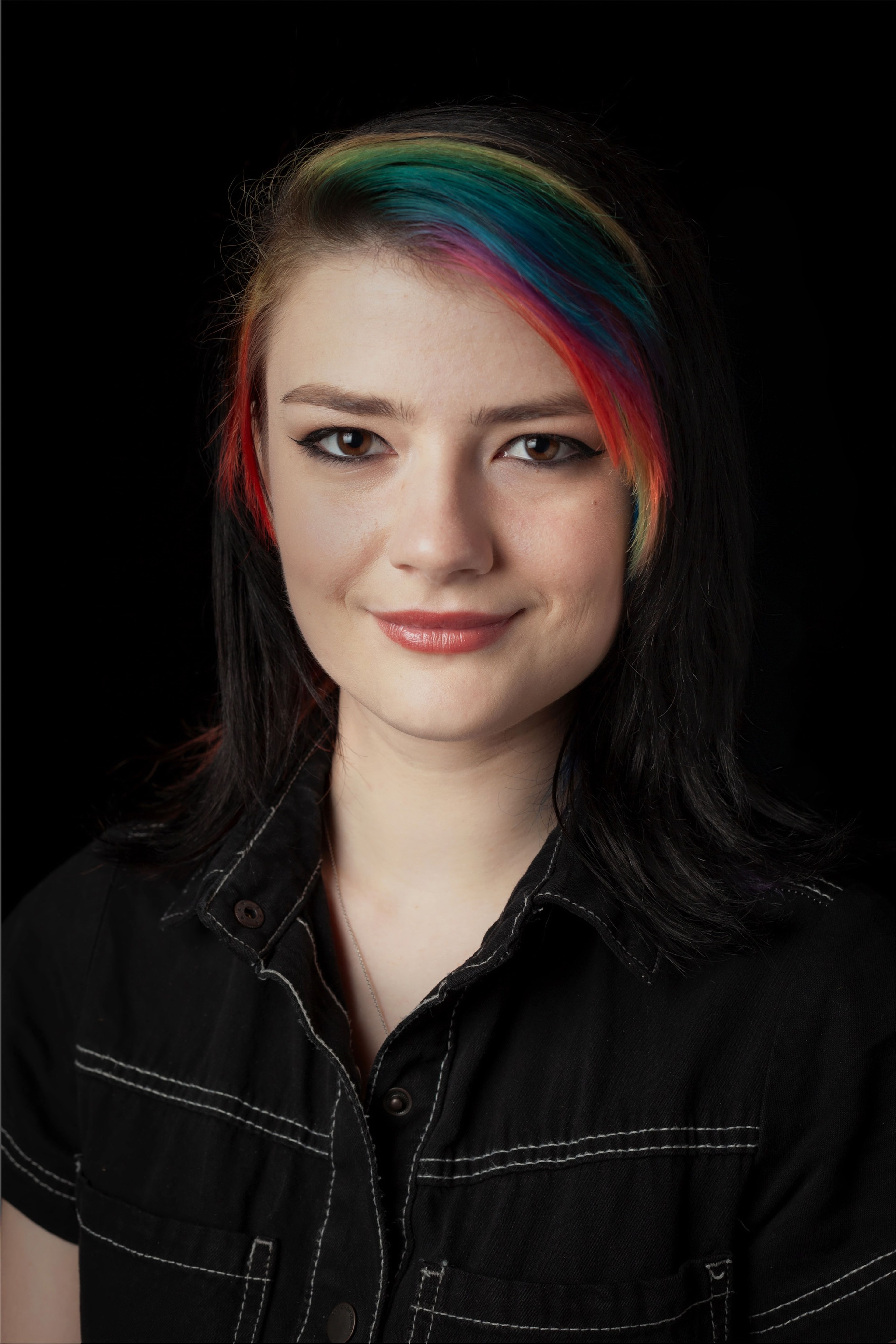 Headshot of a young woman with black hair and vibrant rainbow highlights, wearing a black shirt with white stitching, against a black background.