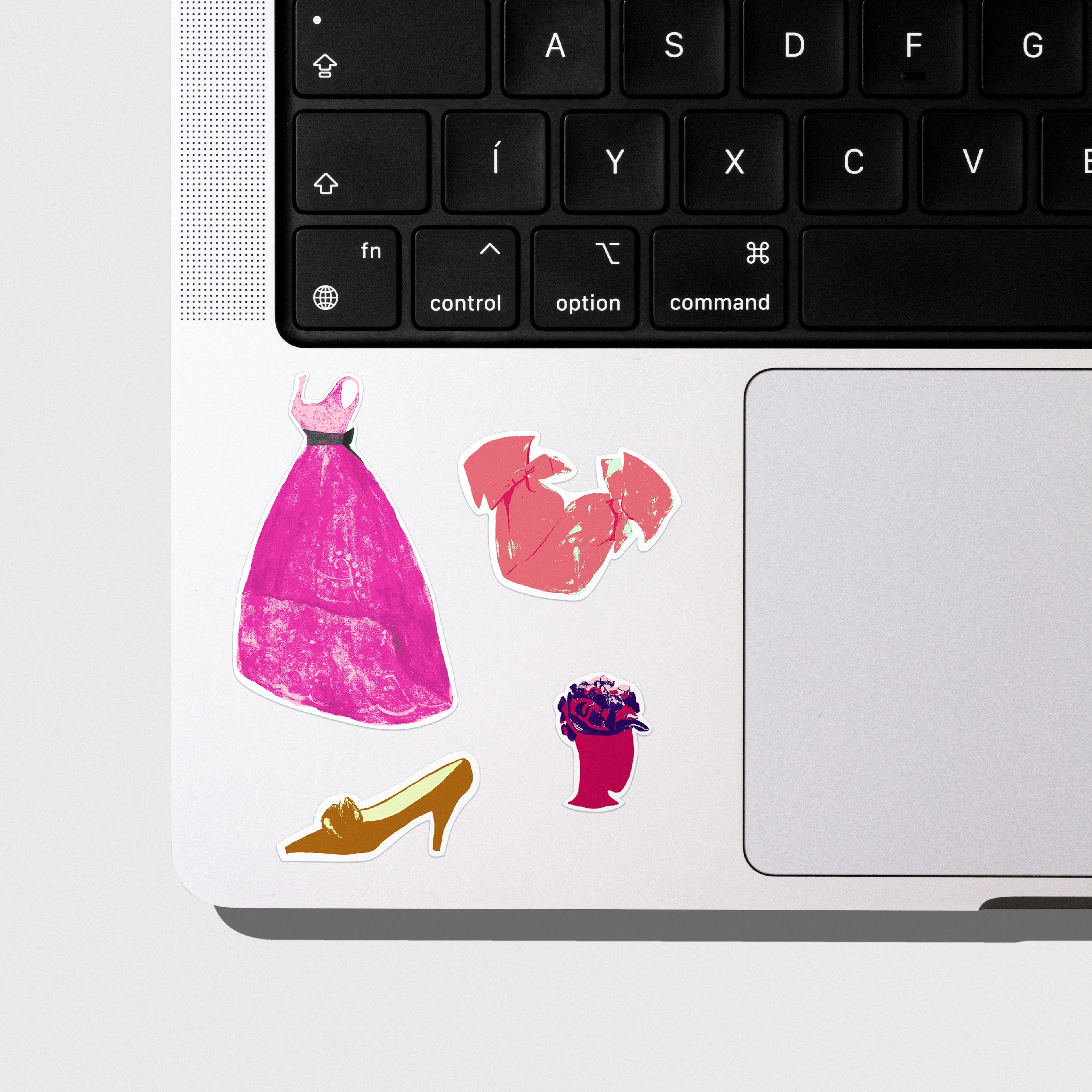 Close-up of stickers featuring fashion items, including a pink dress, yellow high heel, red bow, and stylised hat. The stickers are arranged on the corner of a silver laptop.