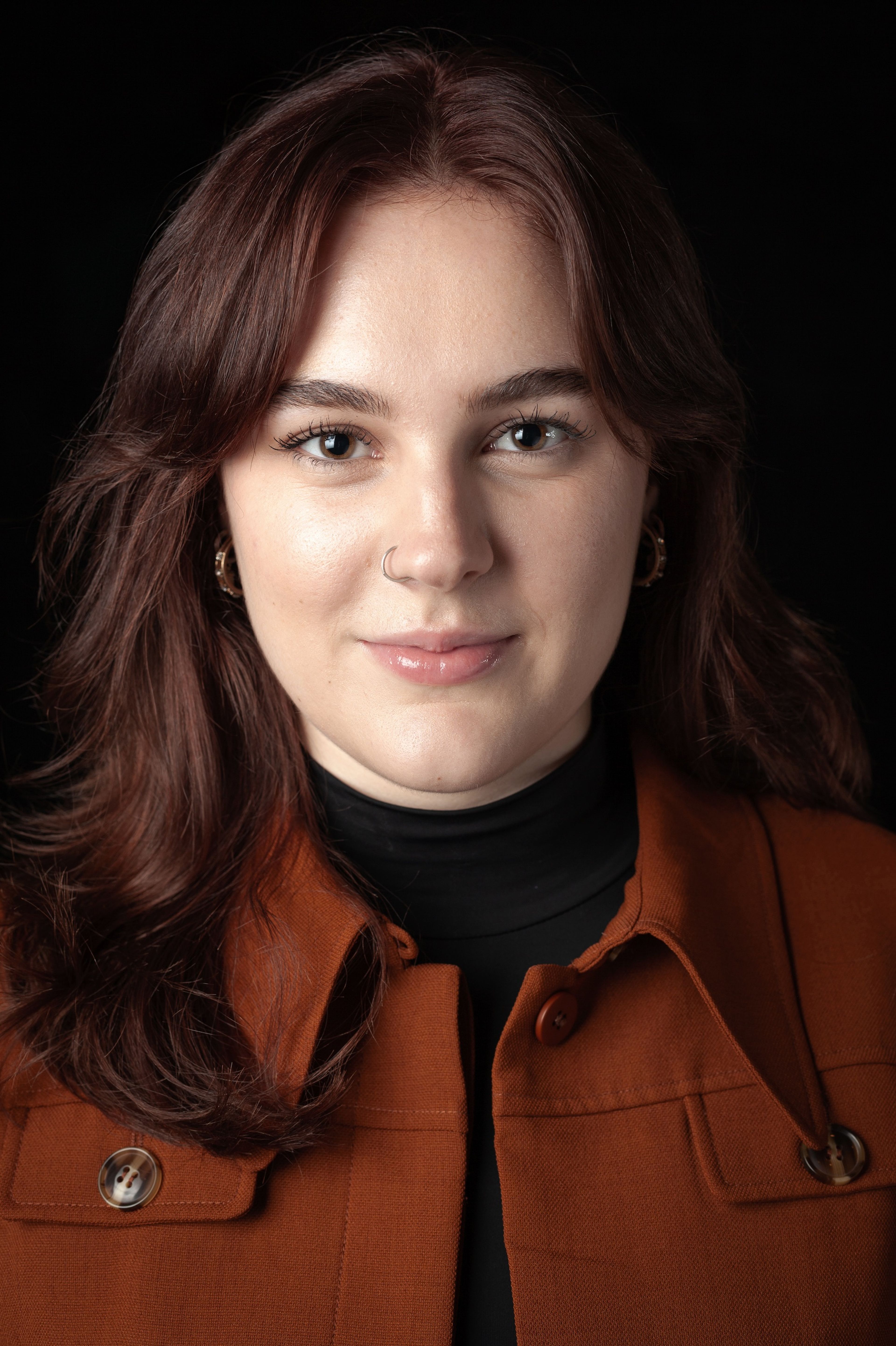 Headshot of a young woman with medium-length auburn hair, wearing a brown coat and black turtleneck, against a black background.