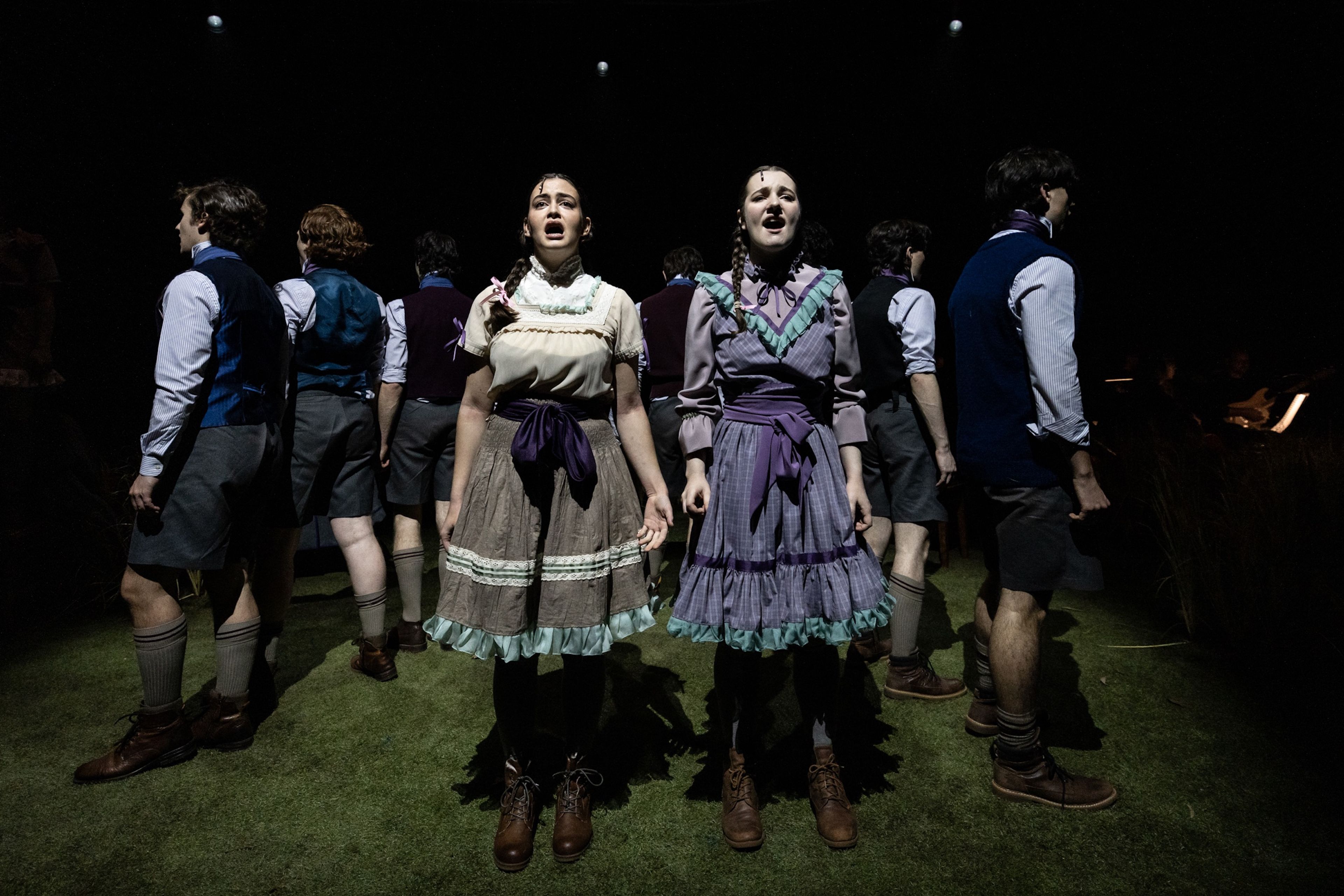 Theatre production photograph showing two performers in ruffled period dresses singing centre stage, flanked by male ensemble members in waistcoats and knee-length trousers, on a dark stage with spotlights above.