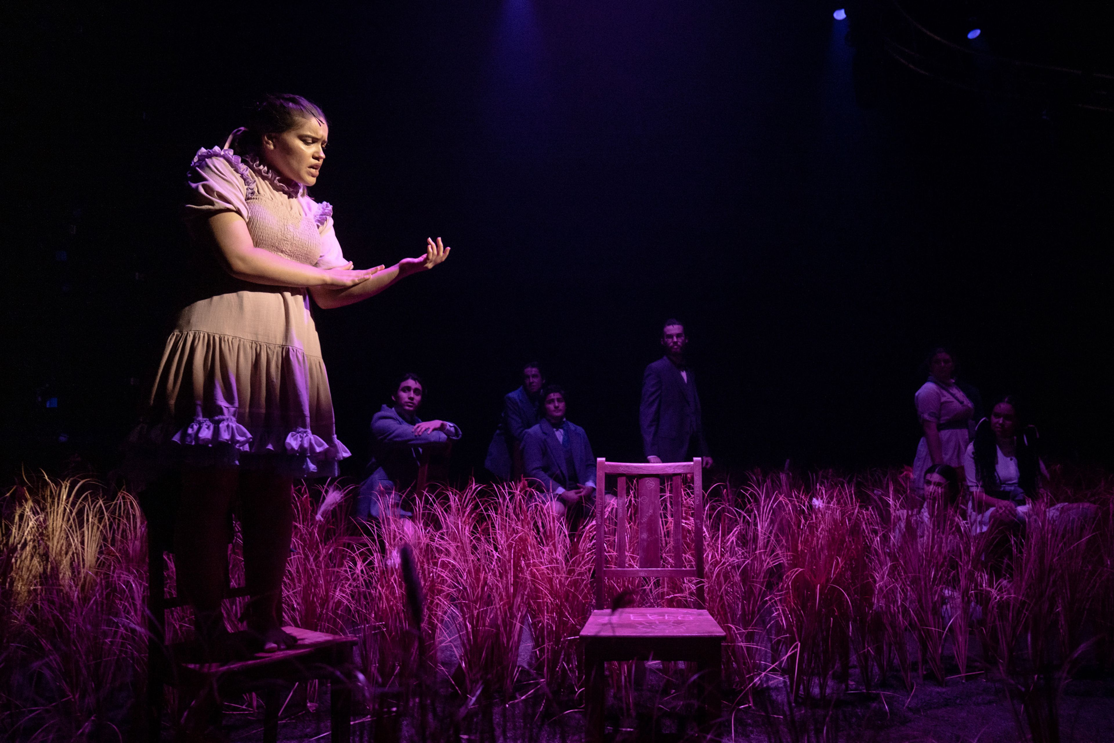 Theatre production photograph of a performer in a Victorian dress standing on a chair delivering a monologue, with other cast members watching from behind wheat grass scenery under purple lighting.