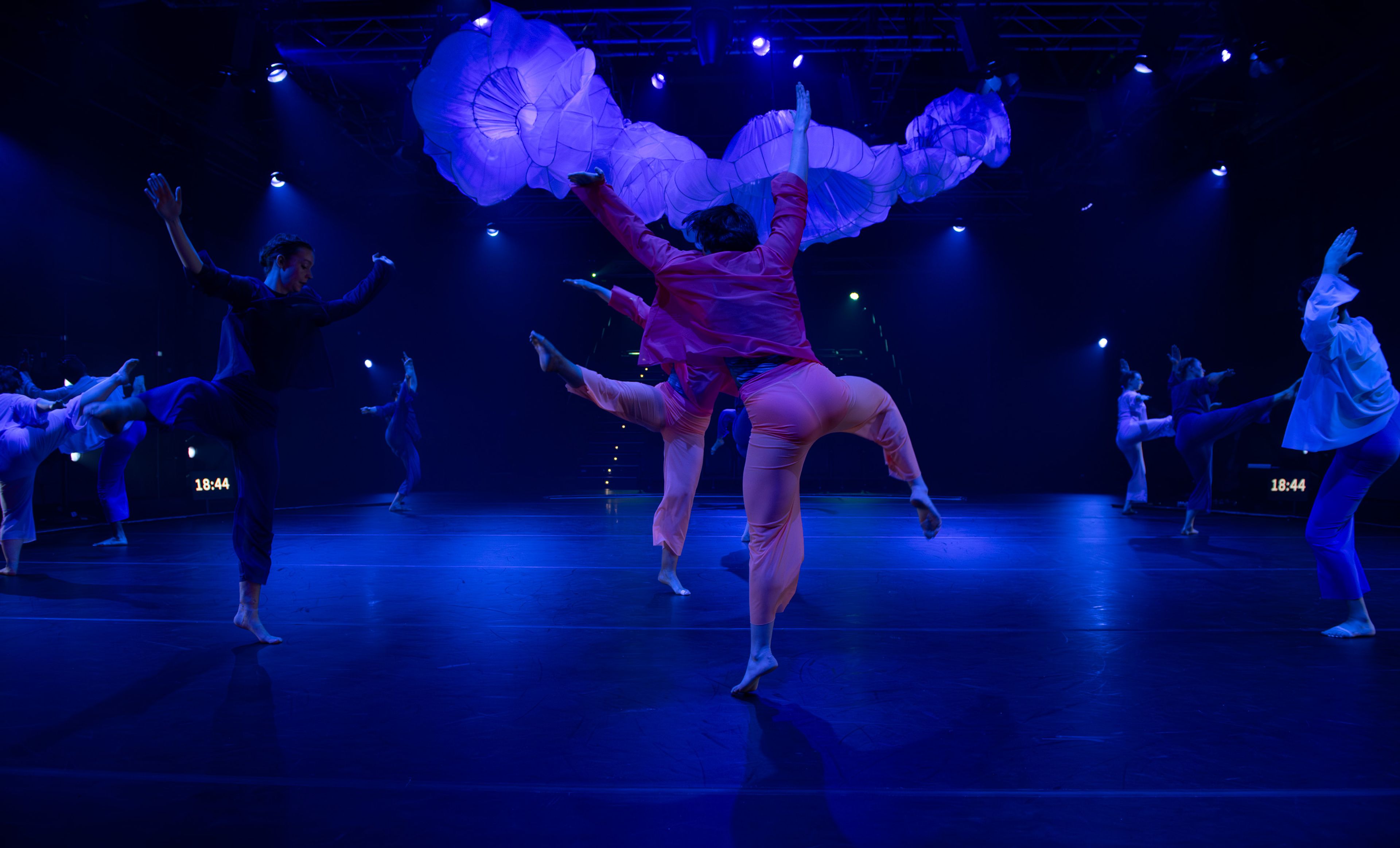 Theatre production photograph with dancers in colourful costumes executing high kicks in synchronised formation on stage, with large, white fabric structures overhead and deep blue lighting.