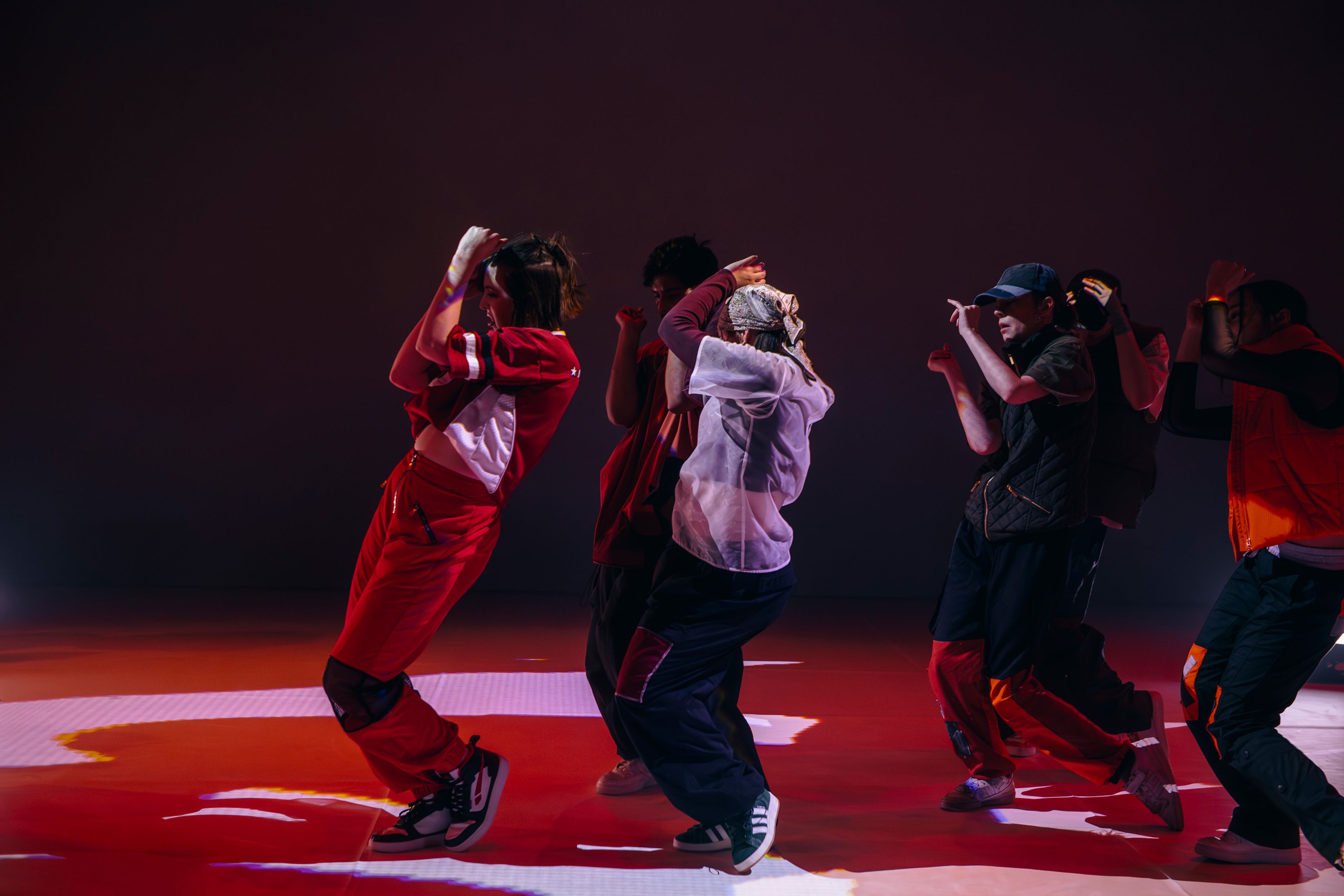 Dance production photograph showing dancers moving together in close formation, lit with strong red and lavender tones.