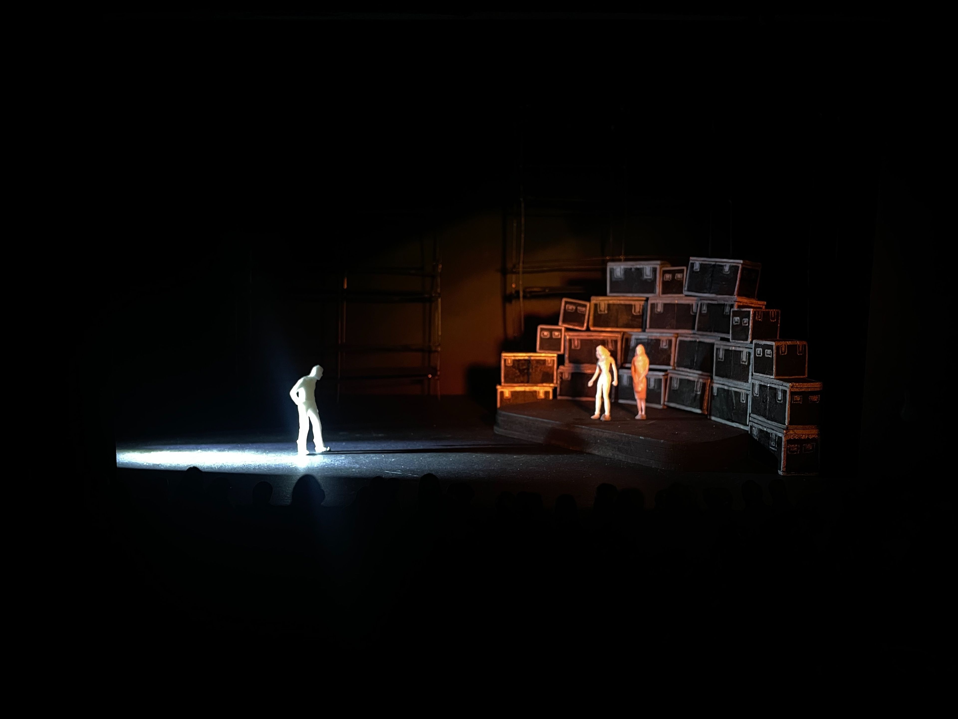 Scale model of a set design, showing a lone figure in spotlight and a stack of stage cases against a dark backdrop.