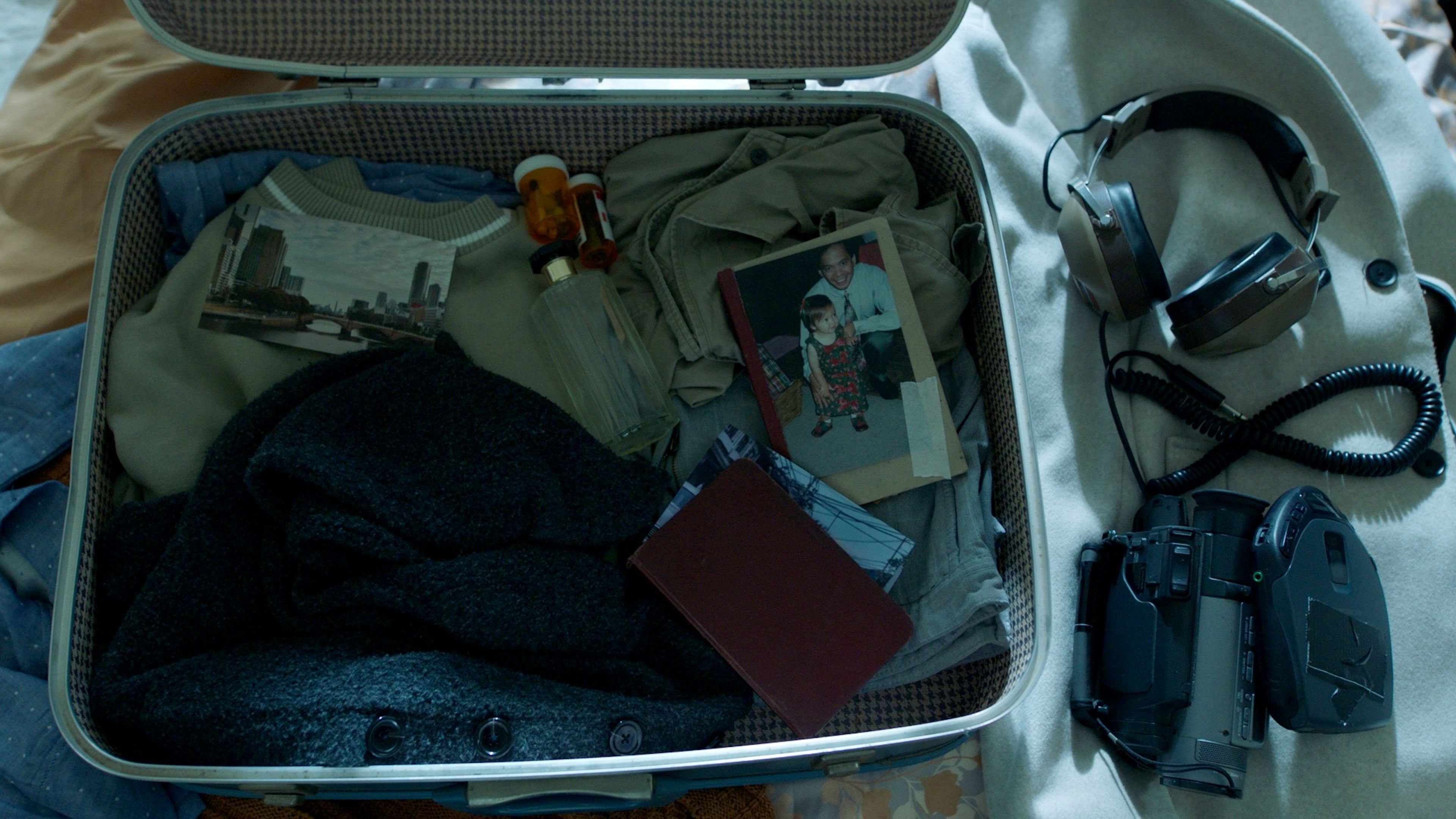 Film prop still life of an open suitcase containing vintage clothing, old photographs, and memorabilia, with headphones and camera equipment beside it