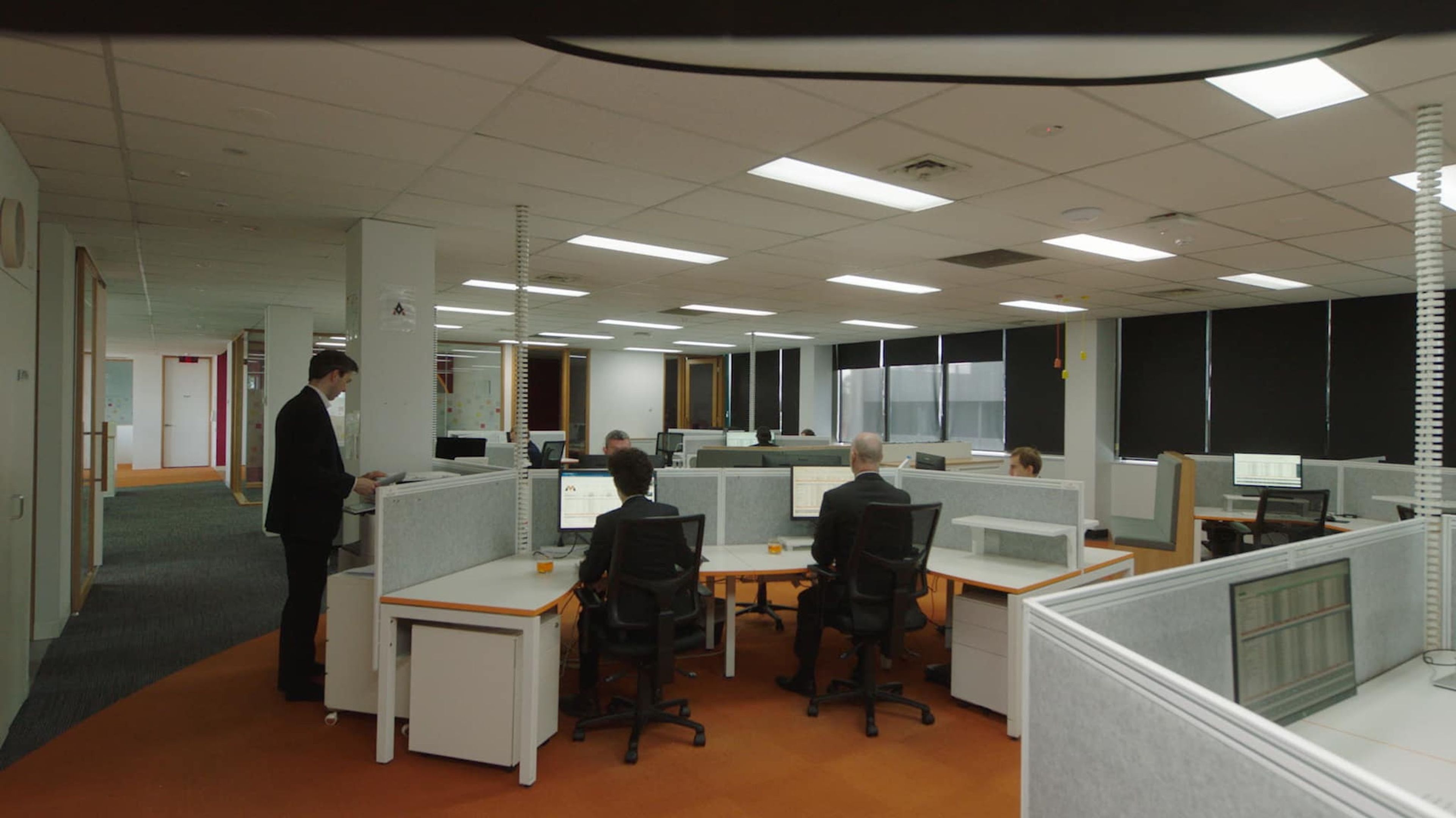 Film still of an open-plan office setting with workers seated at individual cubicles, typing on computers.