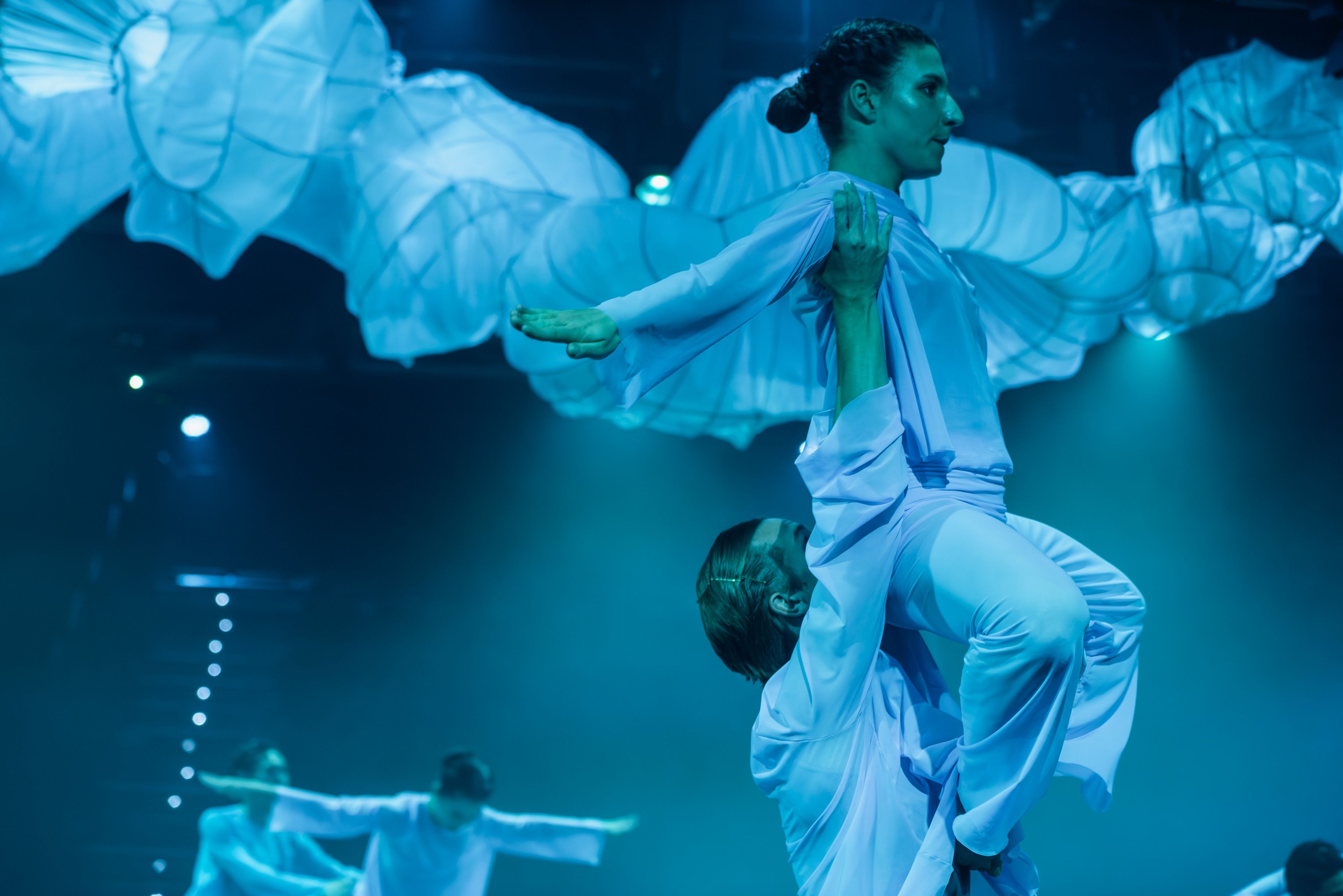 Theatre production photograph showing two dancers in flowing white costumes performing a lift sequence, with sculptural fabric forms visible overhead bathed in turquoise-coloured light.