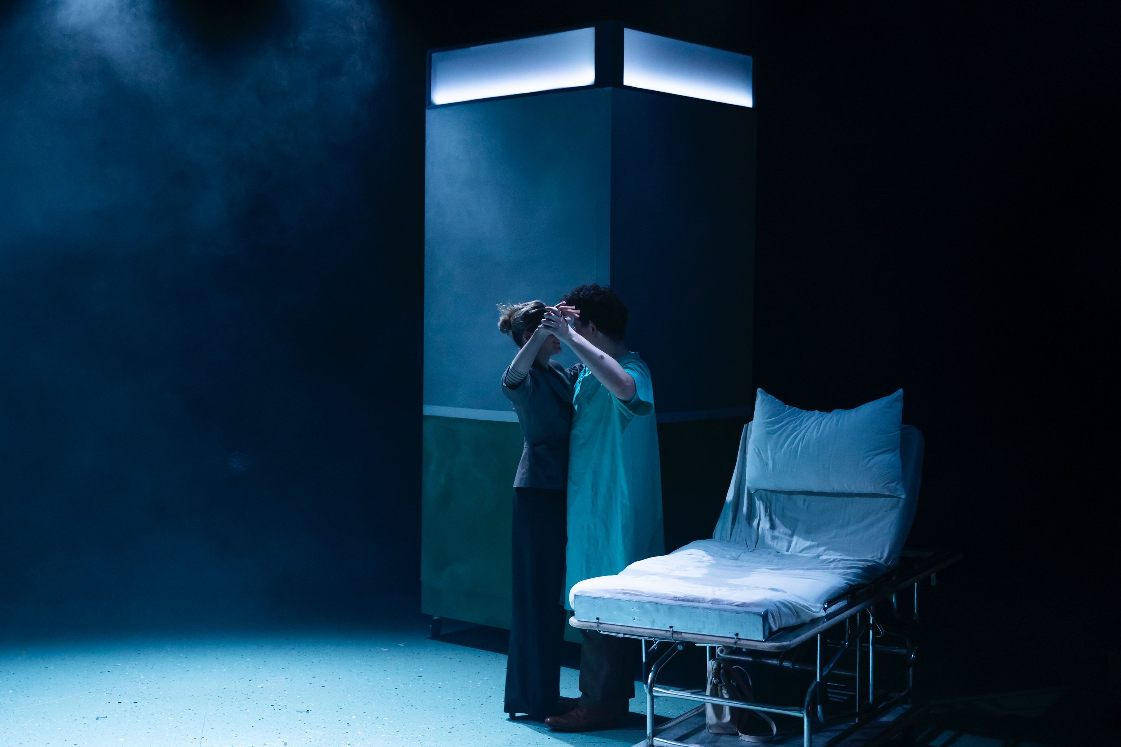 A medical scene with bright overhead lighting, featuring a hospital bed and two performers in front of an illuminated pillar against a dark blue backdrop.