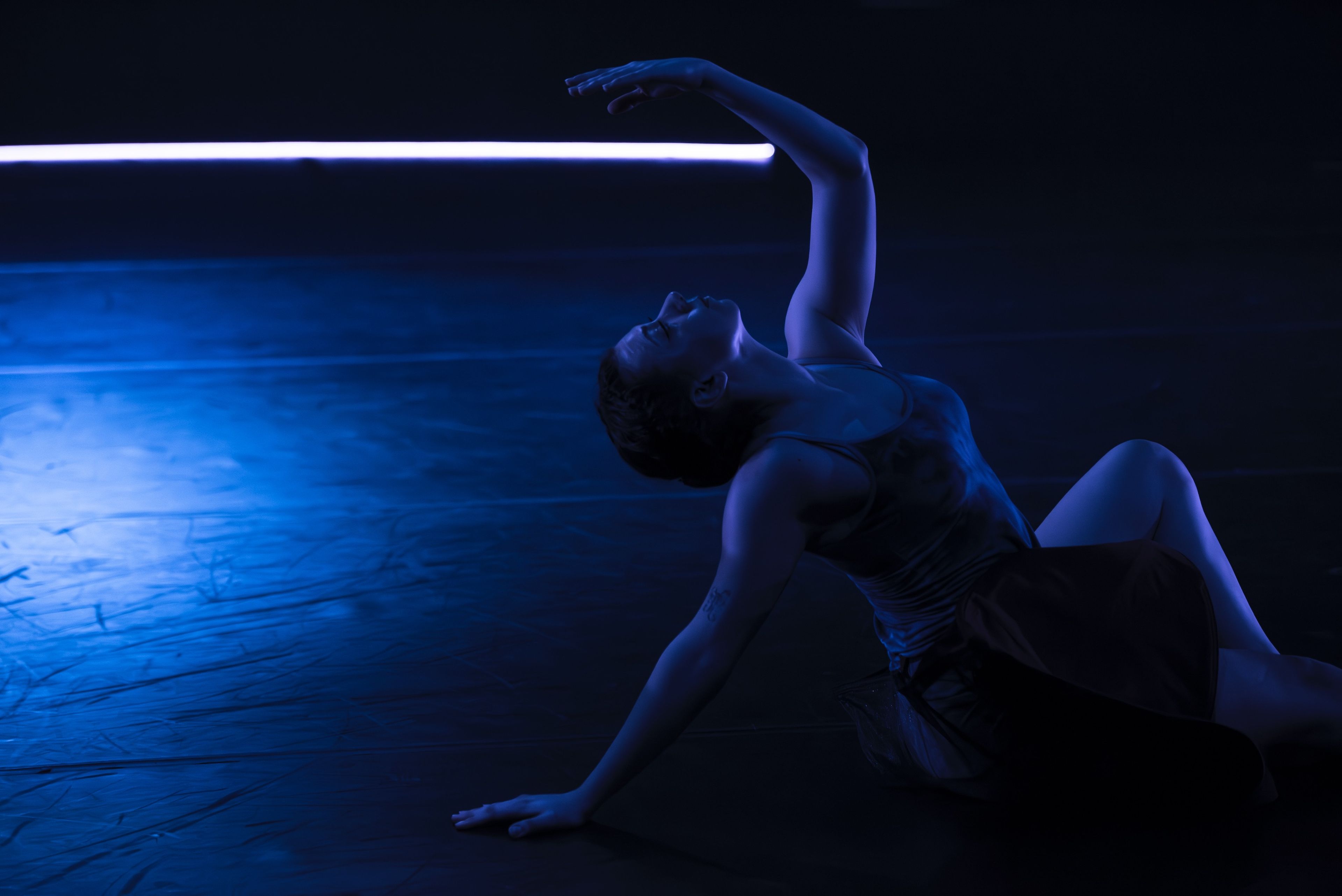 Theatre production photograph of a dancer lying on the stage in a deep blue light, arm extended overhead, creating a sense of fluid movement and expression.