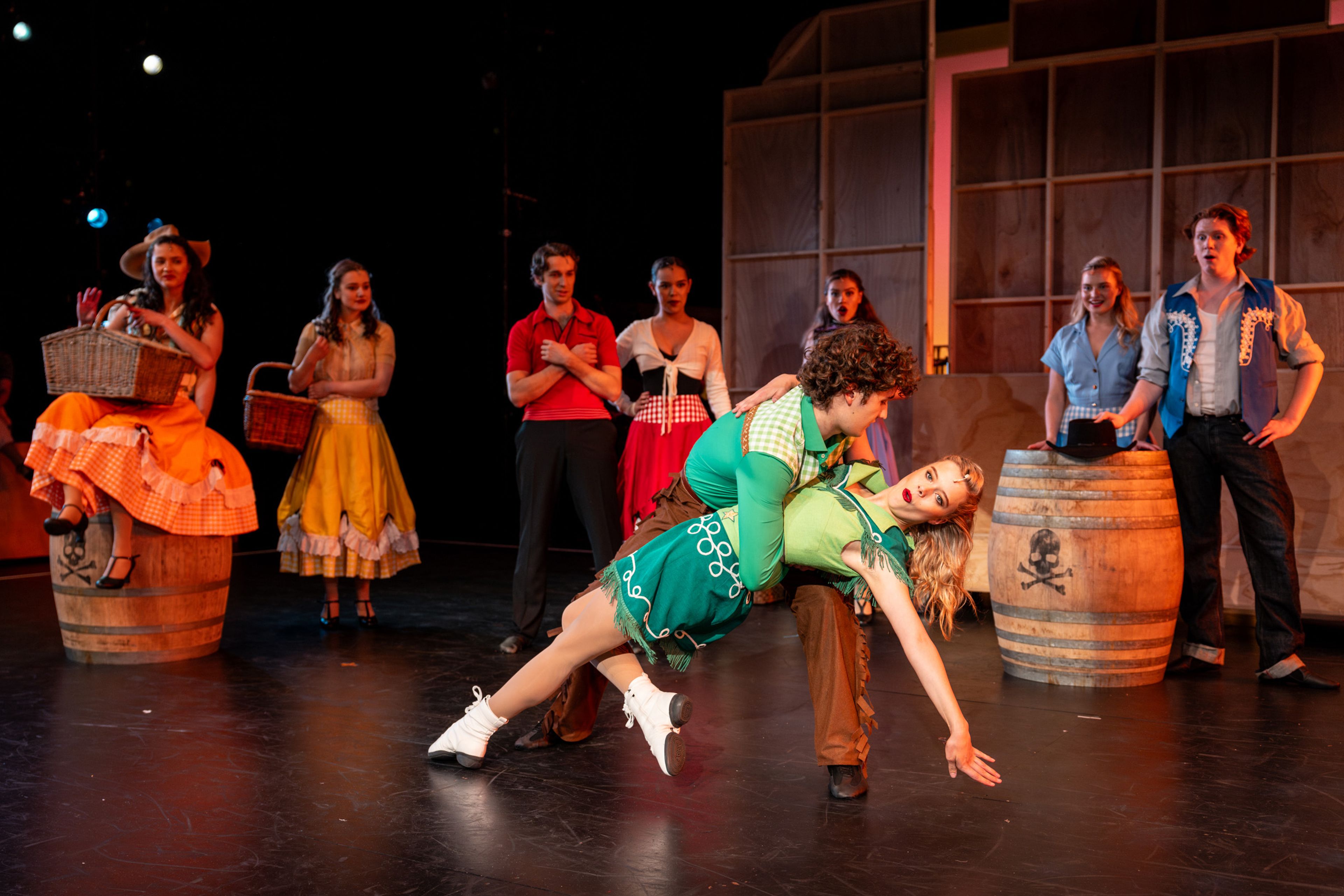 Theatre production photograph showing a dance scene with rustic barrel props, featuring two performers in green costumes in a dramatic dance move while others watch.