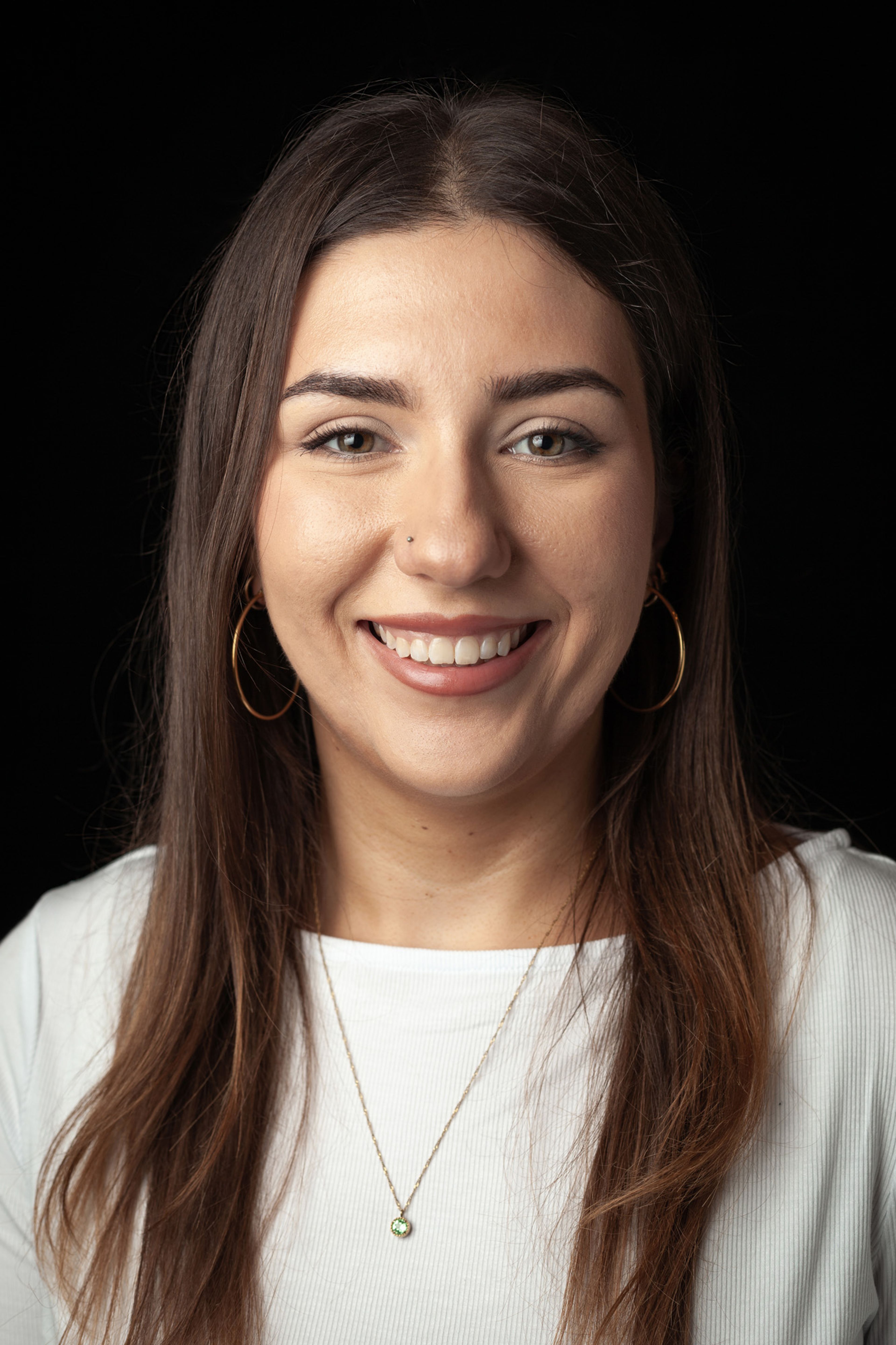 Headshot of a young woman in a white top against a black background, wearing gold hoop earrings and a pendant necklace, with long brown hair and a warm smile.