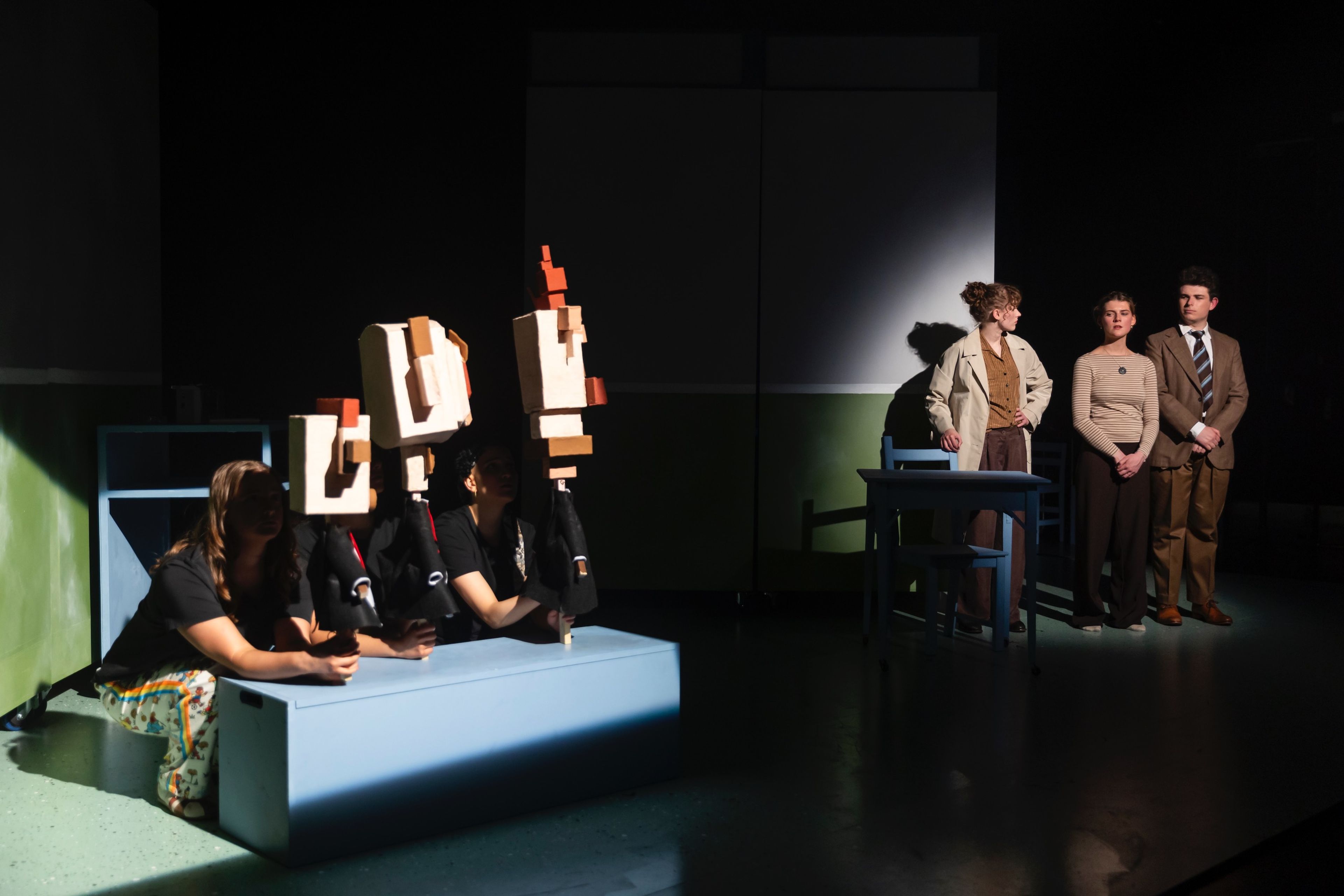 Theatre production photograph with actors holding abstract blocky puppets, partially illuminated on stage, while three others stand in shadow observing.