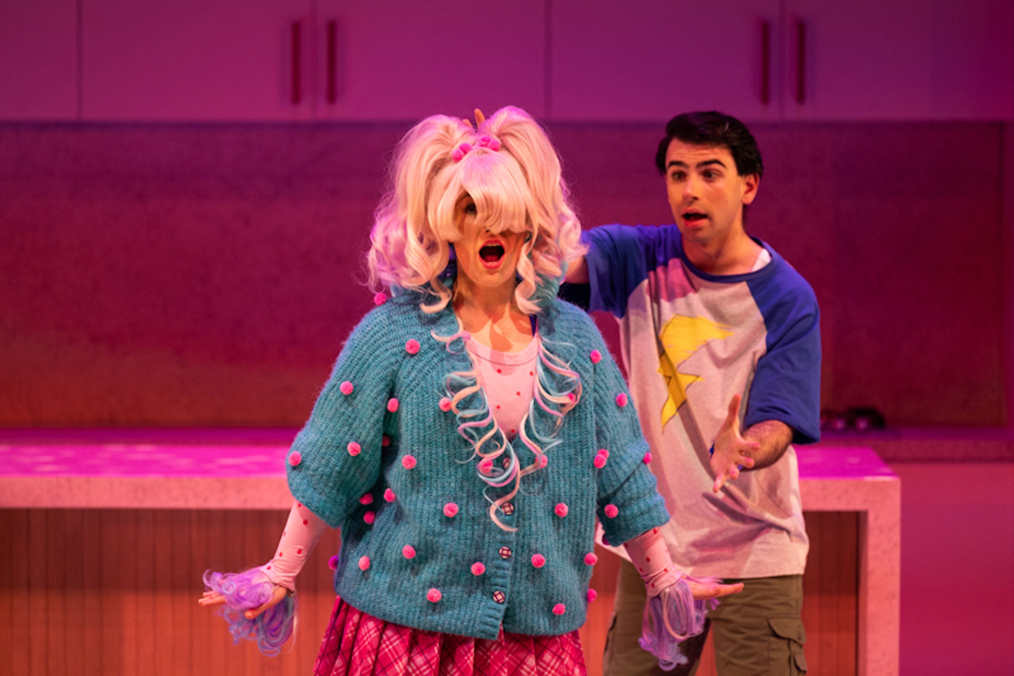 Production photograph featuring two performers under pink lighting - one wearing an extravagant blonde wig, turquoise cardigan with pink pom-poms, and the other in a baseball-style t-shirt