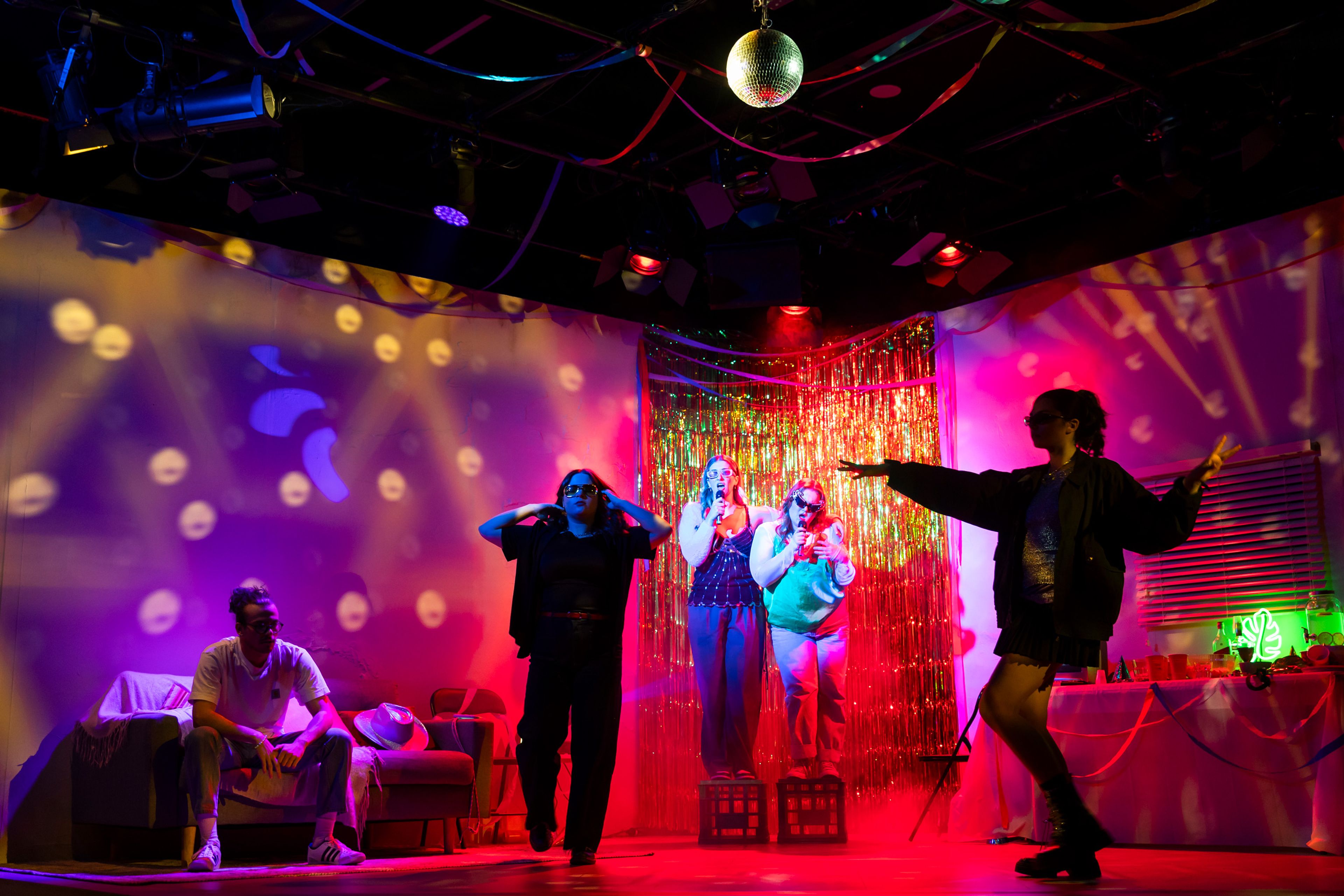 Theatre production photograph capturing a party scene with performers in casual clothes, silver tinsel curtain, and disco ball, lit with vibrant pink and blue lighting and featuring projected patterns on the walls.