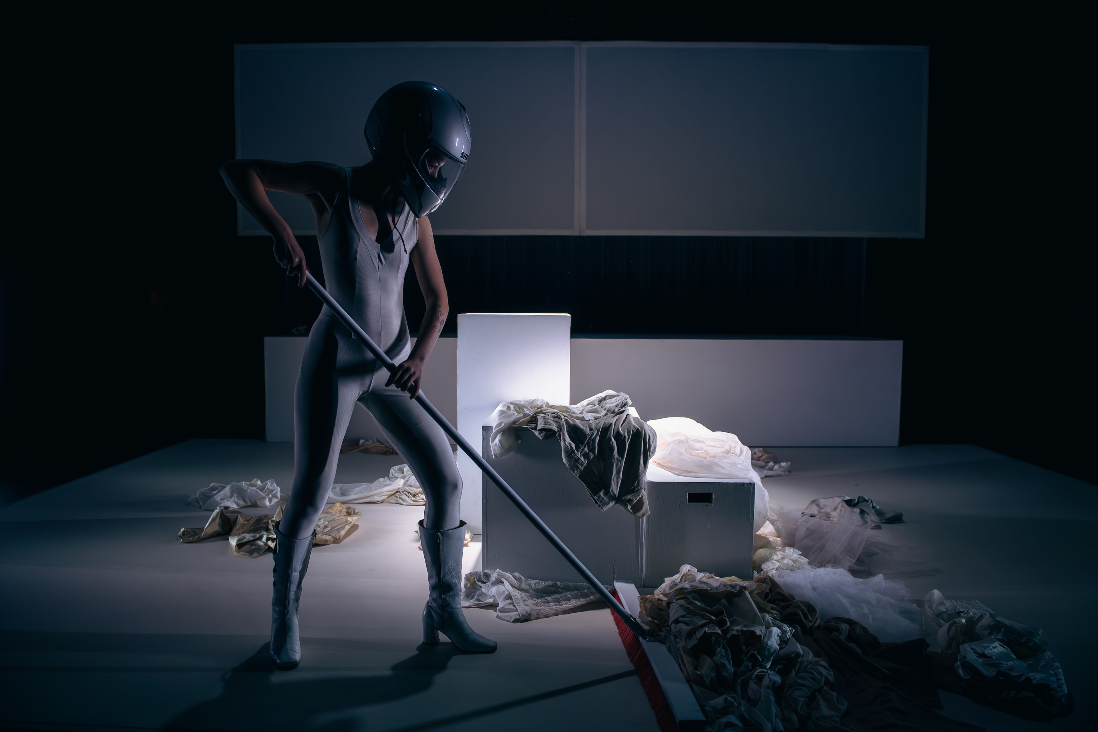 Performance photograph showing a performer in a grey bodysuit and helmet wielding a long pole amidst scattered white fabric and illuminated boxes on a minimalist set.