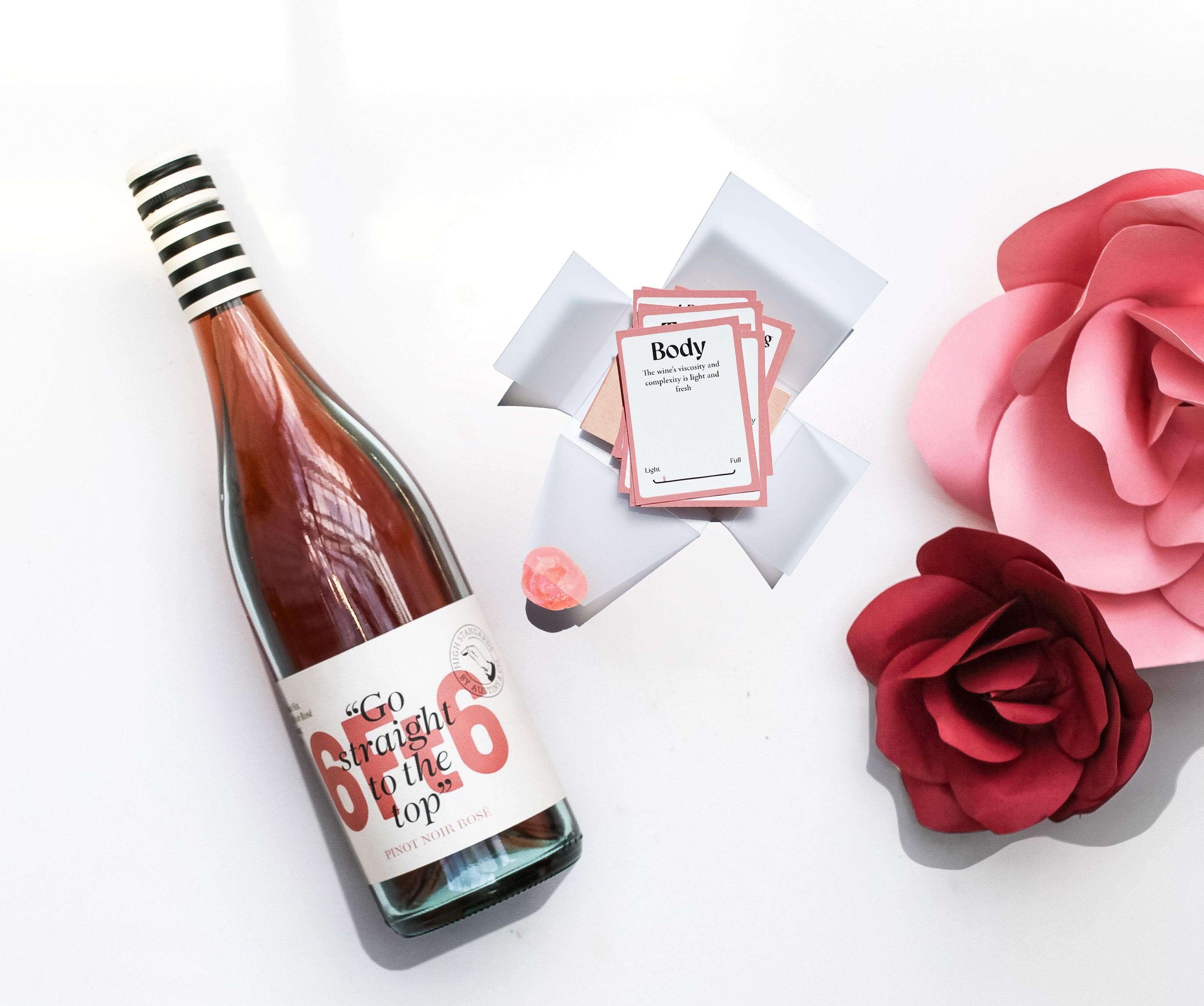 Product styling featuring a Pinot Noir rosé bottle with striped cap, accompanied by wine tasting cards and paper roses in shades of pink against white background.